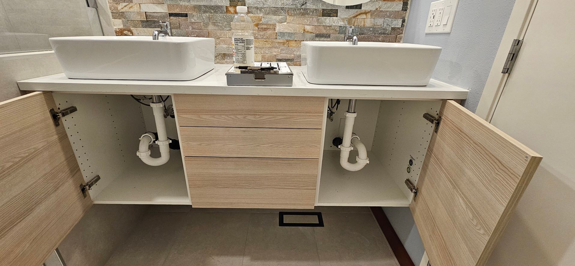 A double bathroom vanity with two white vessel sinks, light wood-toned cabinets, and exposed white plumbing underneath.