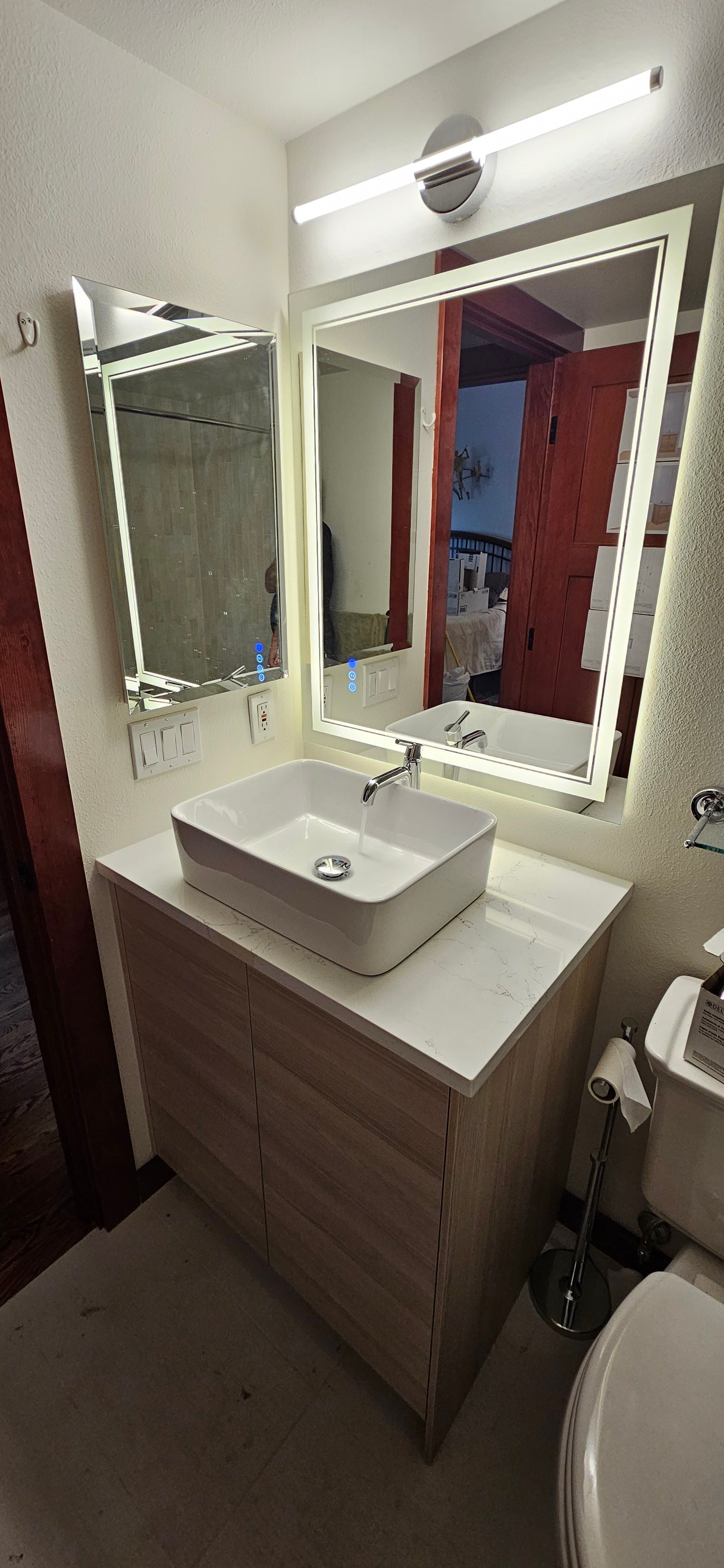 A modern bathroom vanity featuring a white vessel sink on a light wood cabinet with an illuminated mirror and light bar.