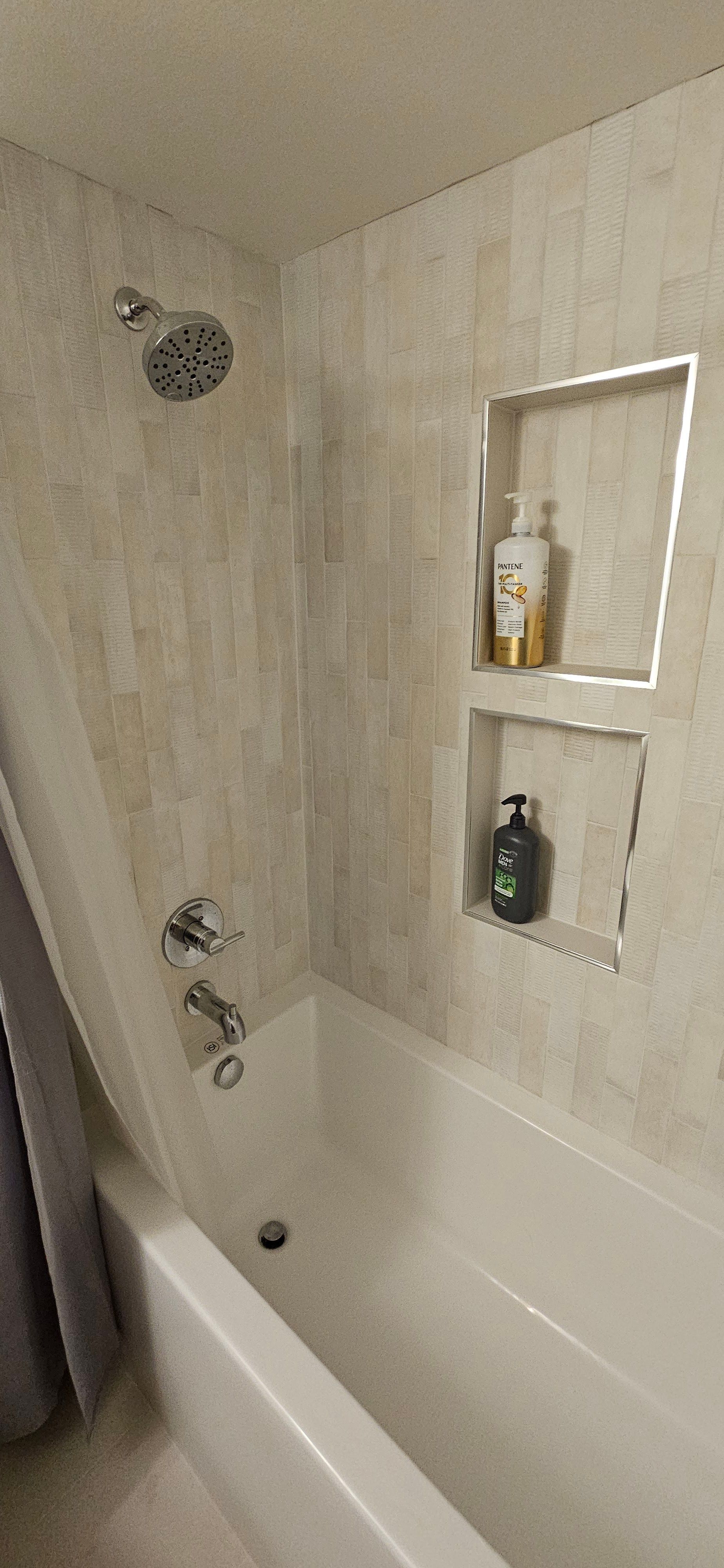 A shower and bathtub enclosure with light-colored tile walls, a chrome shower head, faucet, and a two-tier shower niche.