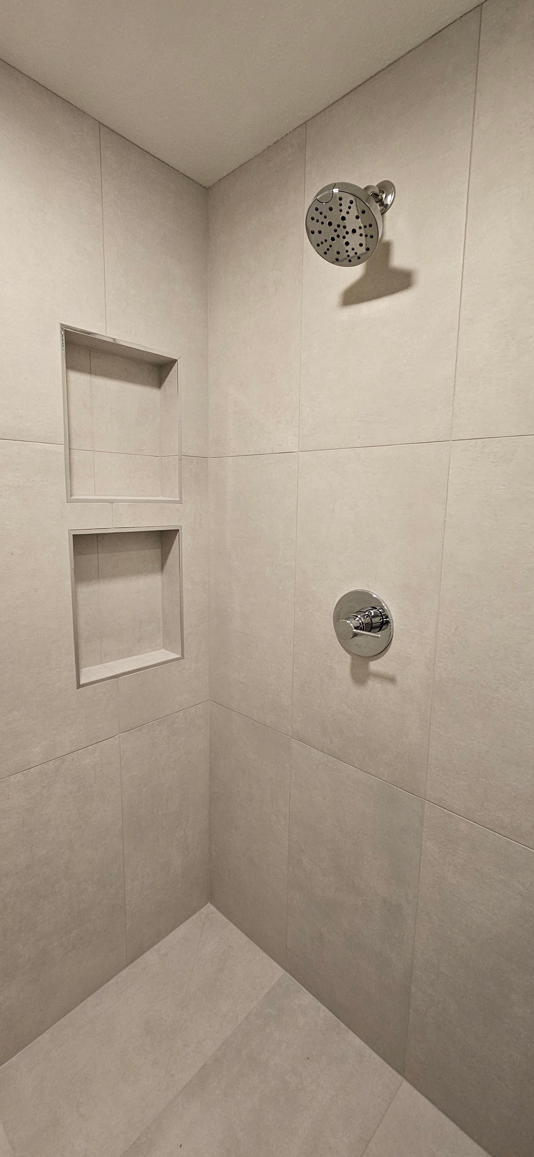 A tiled shower stall with two recessed wall niches, a chrome shower head, and a circular chrome temperature control valve.