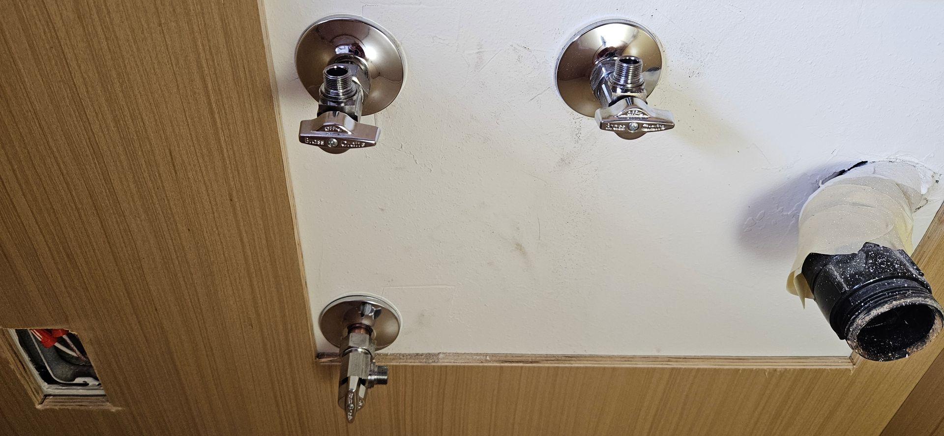 Three chrome plumbing shut-off valves and an open pipe drain stub extending from a white wall inside a wooden cabinet.