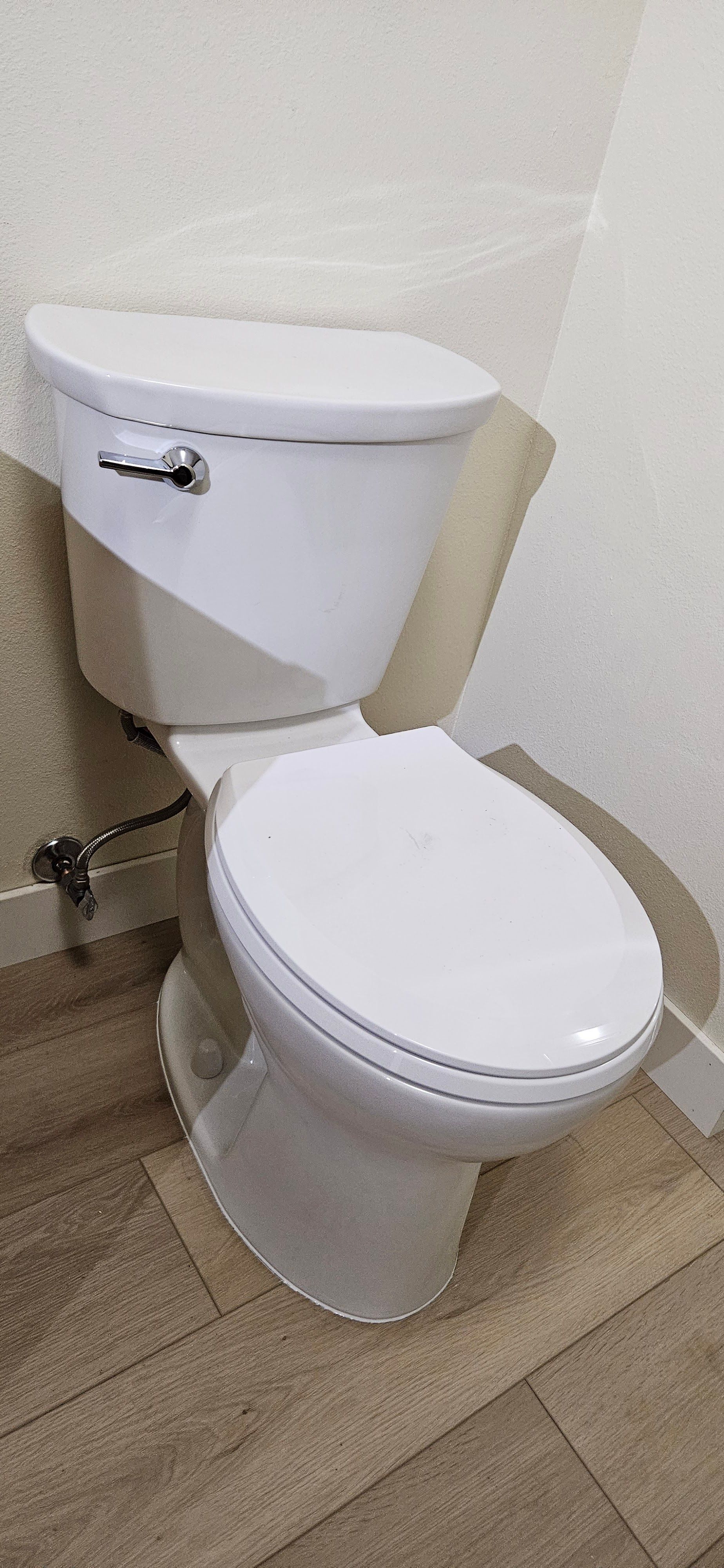 A white porcelain toilet with a closed lid, situated in a bathroom corner with beige tiled flooring and off-white walls.