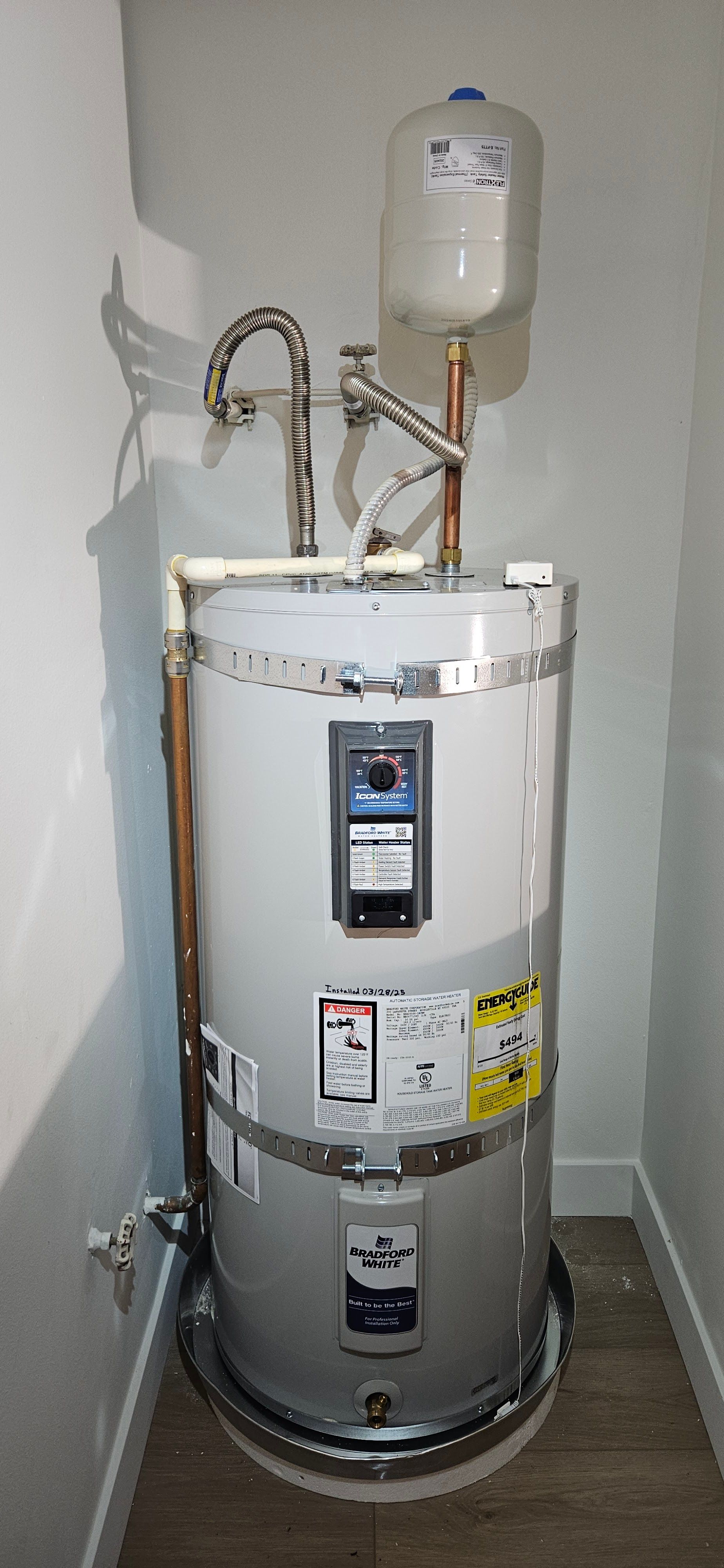 A tall gray electric water heater with an expansion tank, installed in a corner on a white pan and light wood floor.