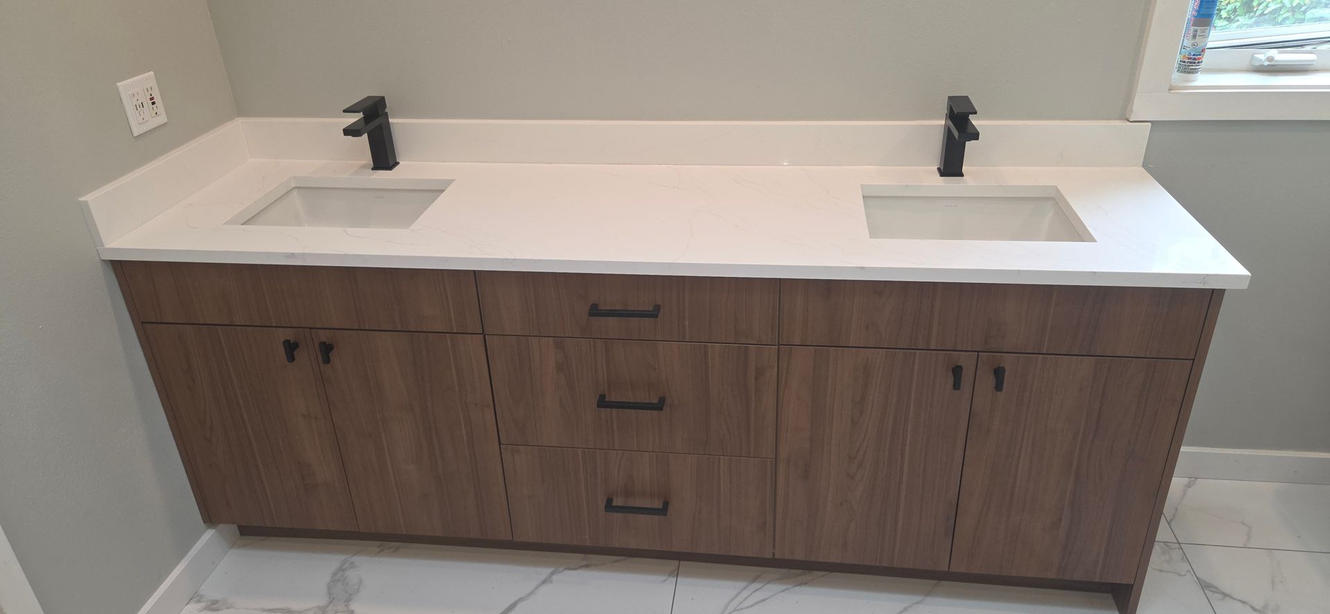 A double-sink bathroom vanity with wood-grain cabinets, black hardware, and a white countertop against a light grey wall.