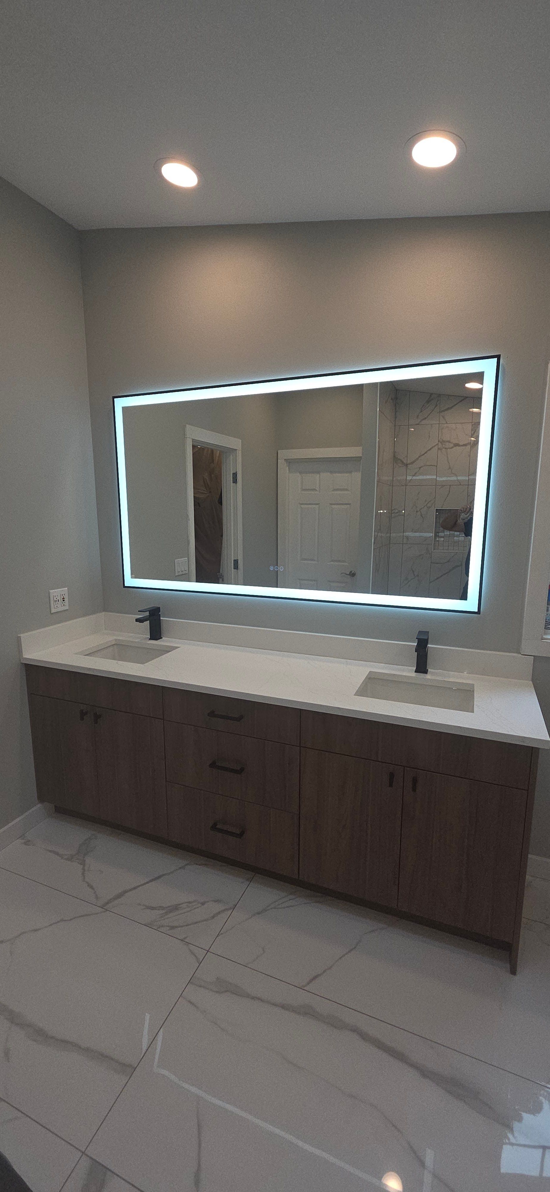 A modern bathroom vanity with a double sink, white countertop, dark wood cabinets, and a large backlit rectangular mirror.