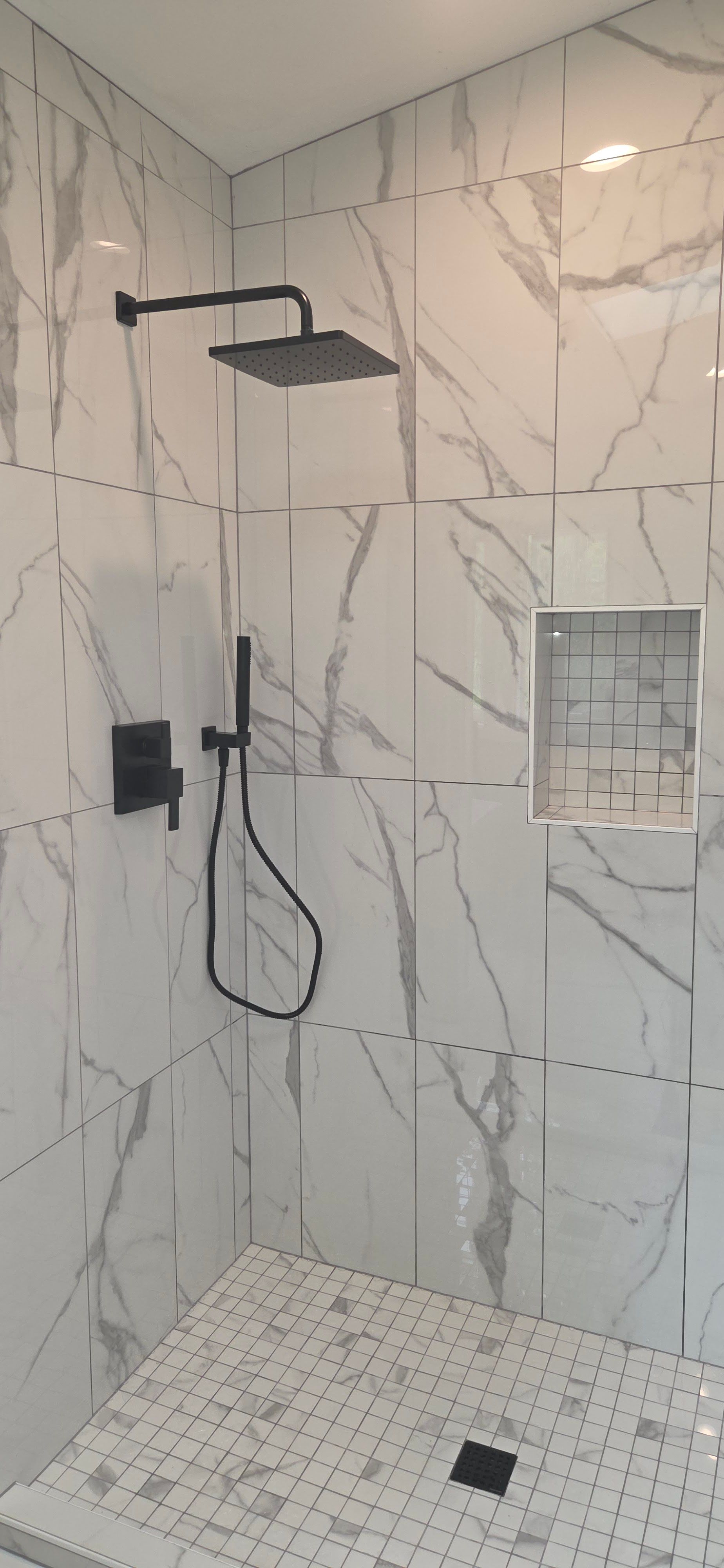A modern walk-in shower with white marble-patterned wall tiles, a black rainfall showerhead, and a mosaic tiled floor.
