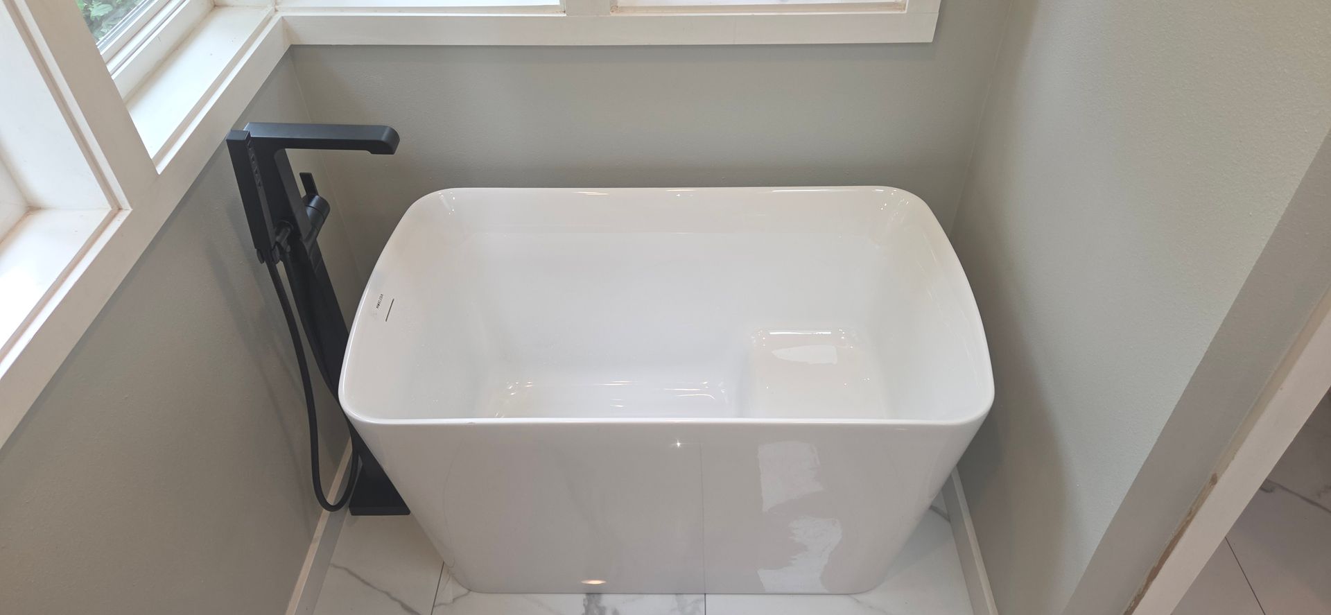 A white freestanding soaking tub in a bathroom corner with a matte black faucet and tiled flooring.