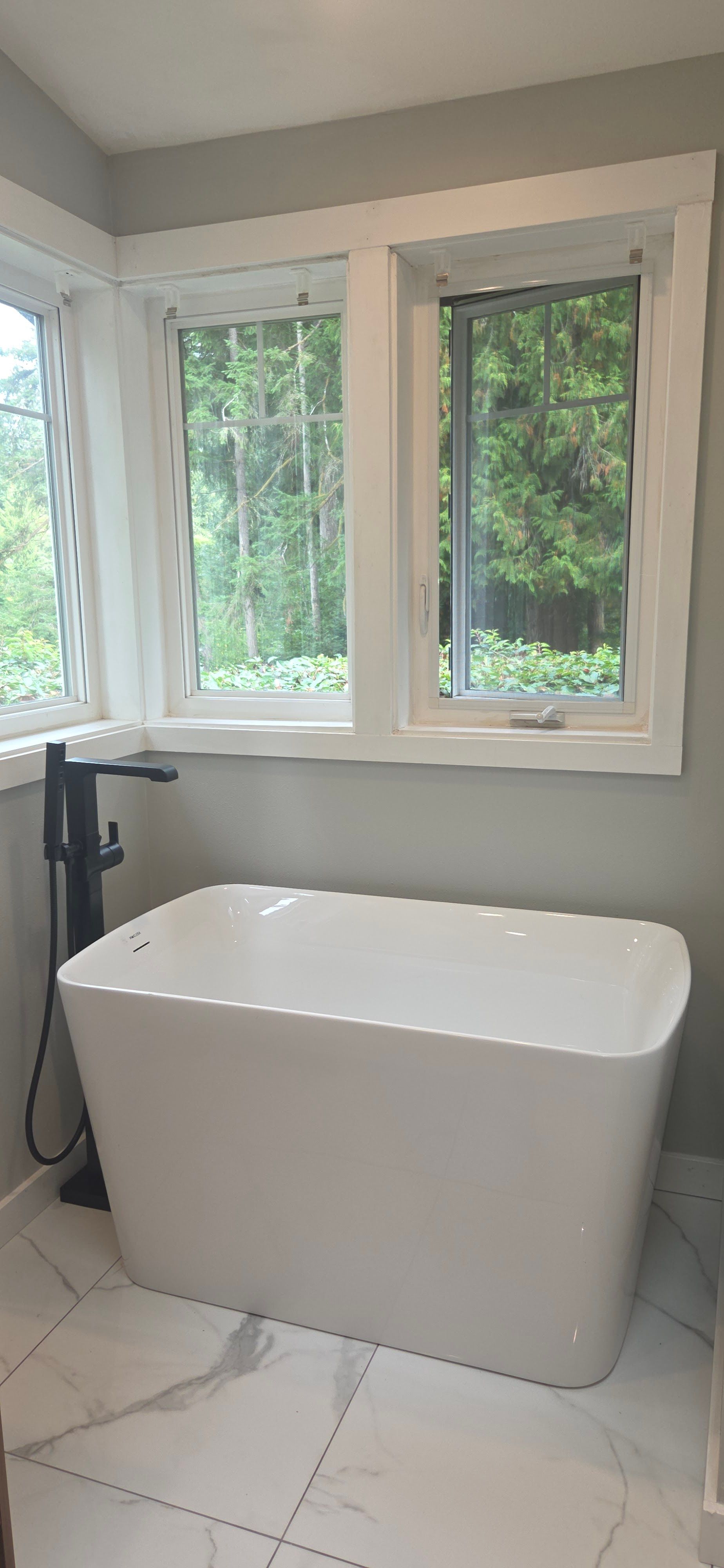 A white freestanding bathtub with a black floor-mounted faucet beneath windows in a tiled bathroom.
