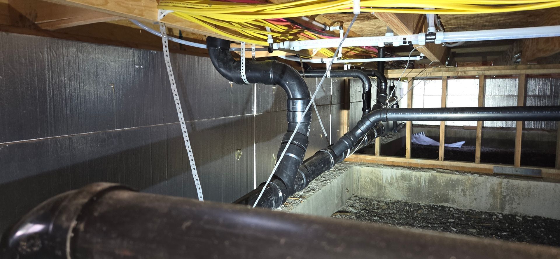 Black plumbing pipes hang from wooden floor joists in a crawlspace with gravel flooring and insulated walls.