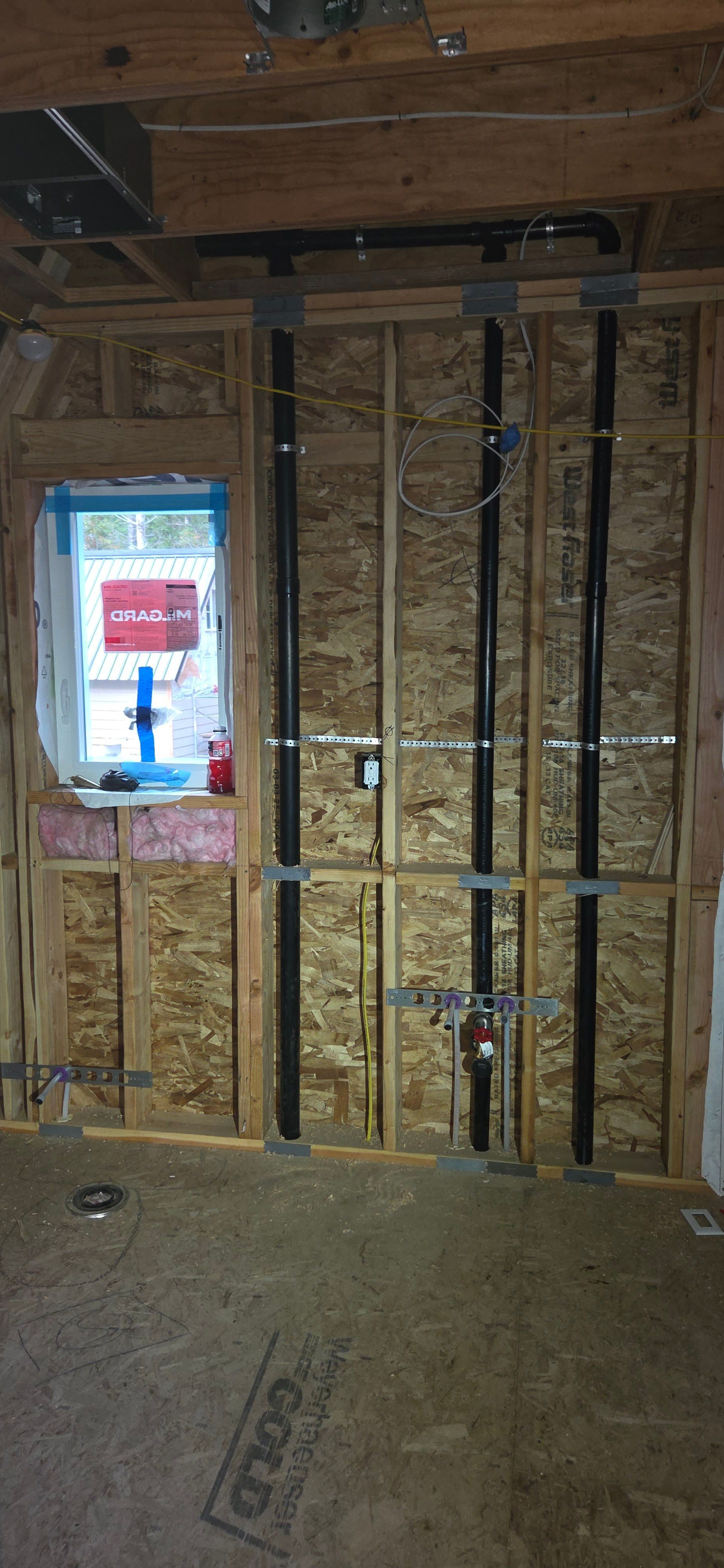 Interior construction view showing exposed wooden wall framing, black plumbing pipes, and a small window under installation.