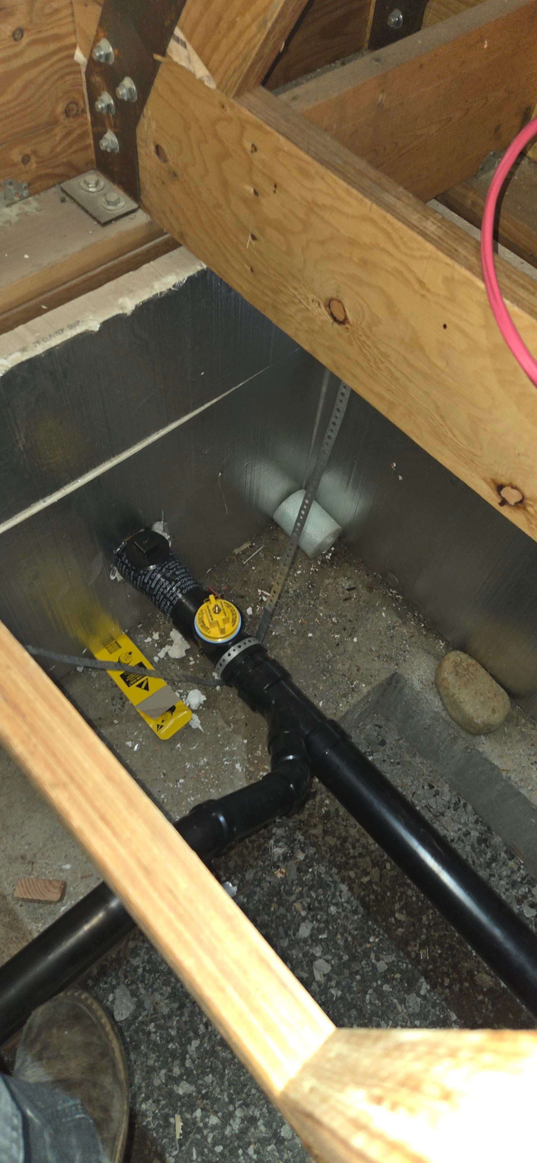 A black plastic pipe with a yellow-handled valve sits in a crawl space under wooden floor joists.