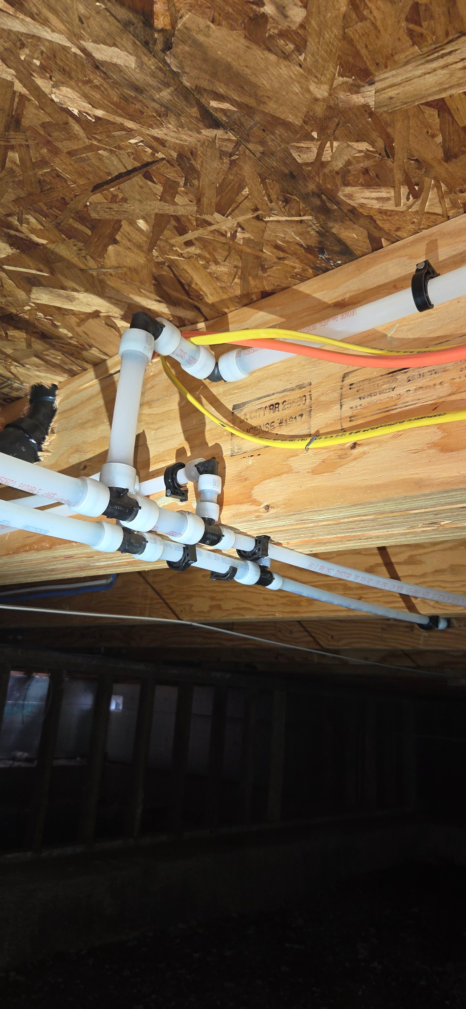 White PEX plumbing lines run horizontally along exposed wooden floor joists, secured with black plastic pipe hangers.