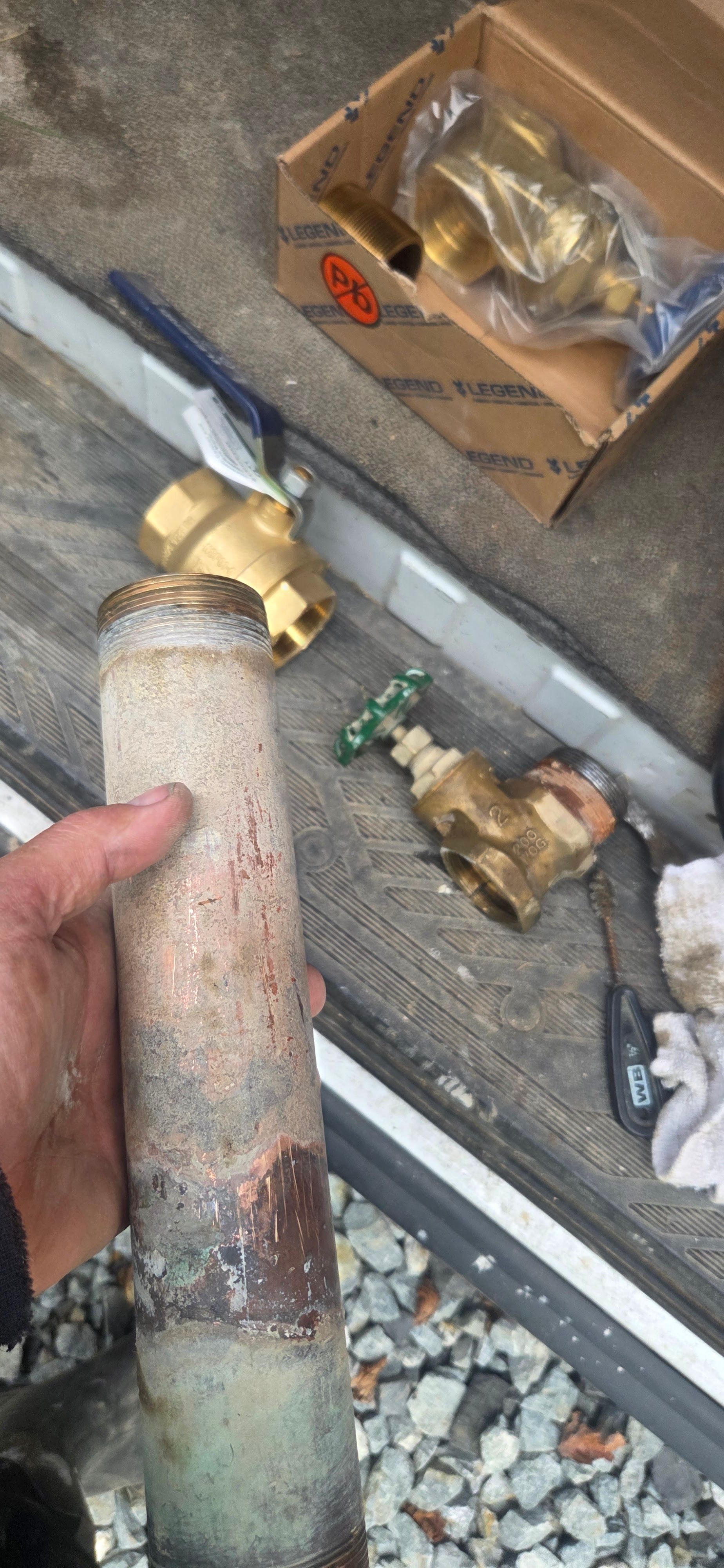 A hand holds a corroded pipe near new brass plumbing valves and parts on a workshop surface.