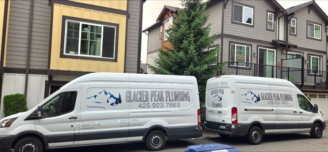 Two white plumber vans parked on a residential street in front of two-story townhouses.