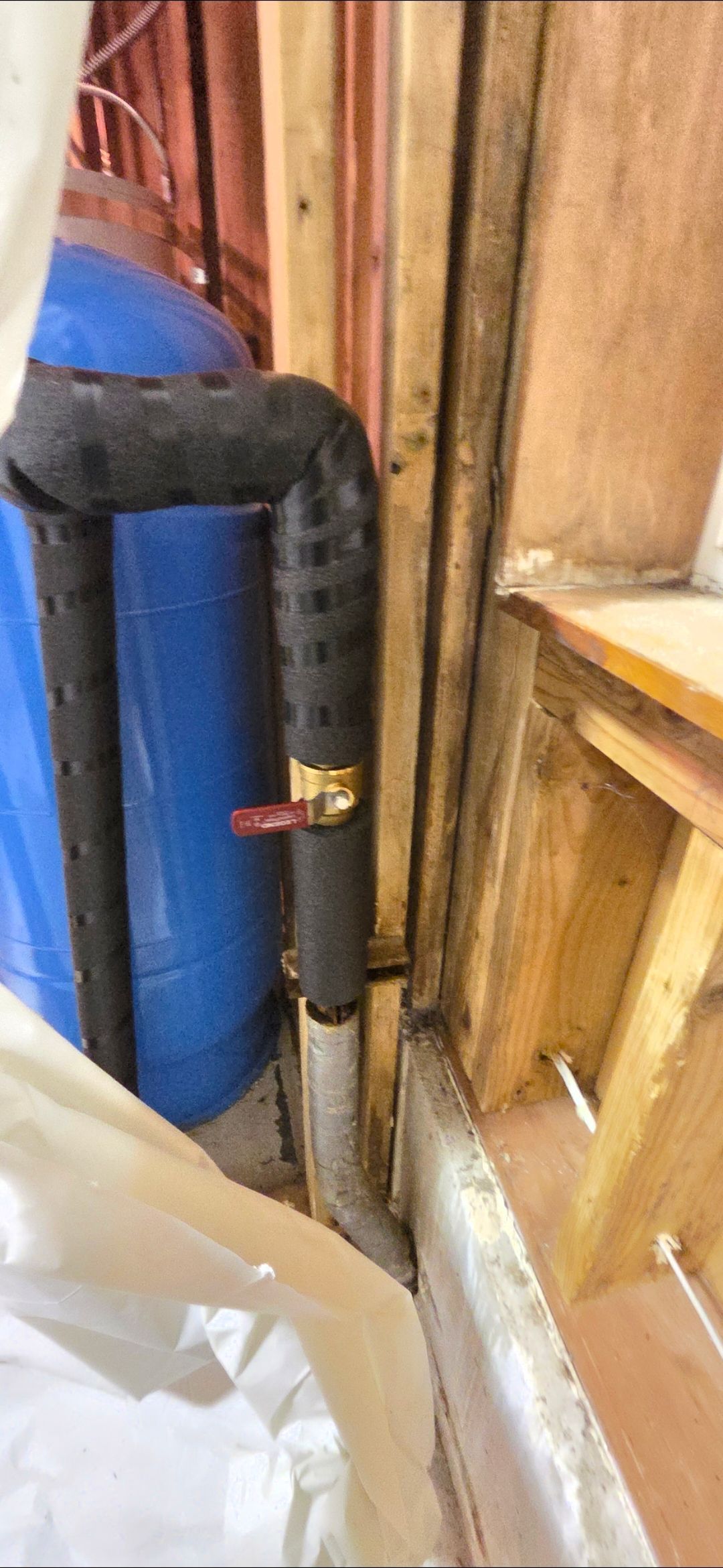 A blue water pressure tank connected to a vertical pipe with a red-handled shut-off valve, framed by wooden wall studs.