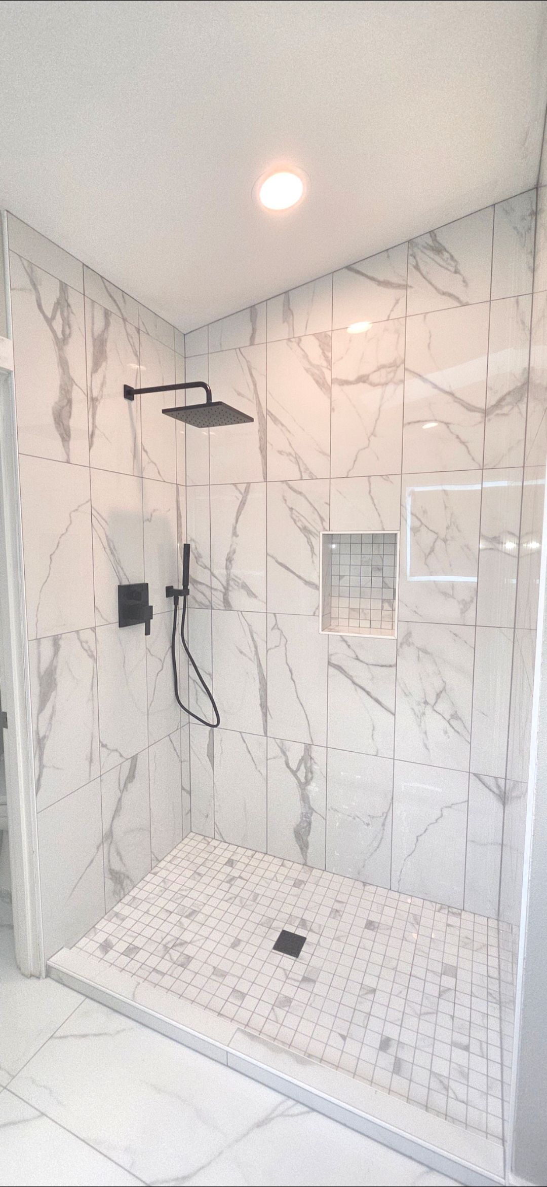 A walk-in shower with marble-style tiled walls, a black rainfall showerhead, handheld sprayer, and a pebble tile floor.