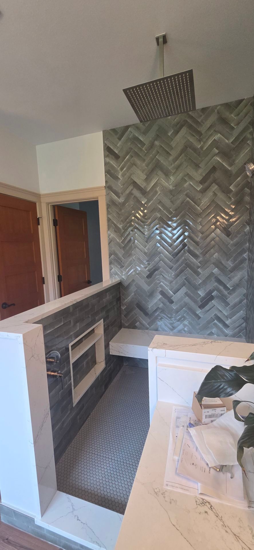 A bathroom shower with gray herringbone-tiled walls, a rainfall showerhead, a floating bench, and a marble vanity counter.