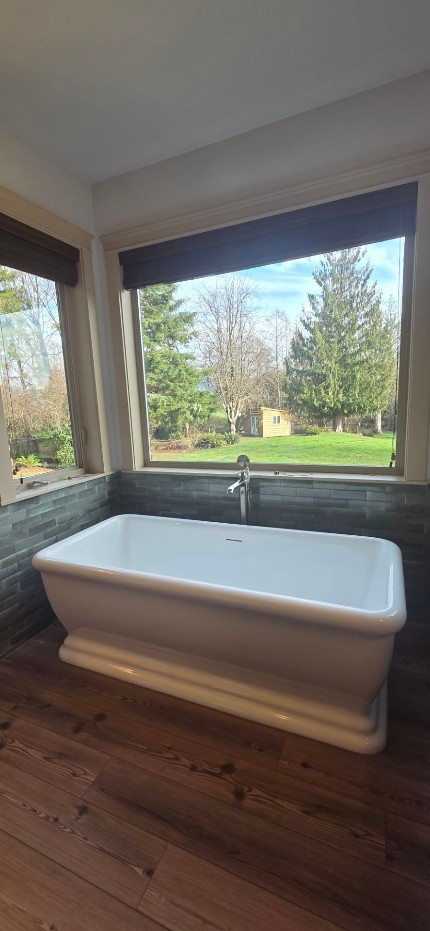 White freestanding bathtub against a slate-tiled wall beneath a large window overlooking a grassy yard with trees.