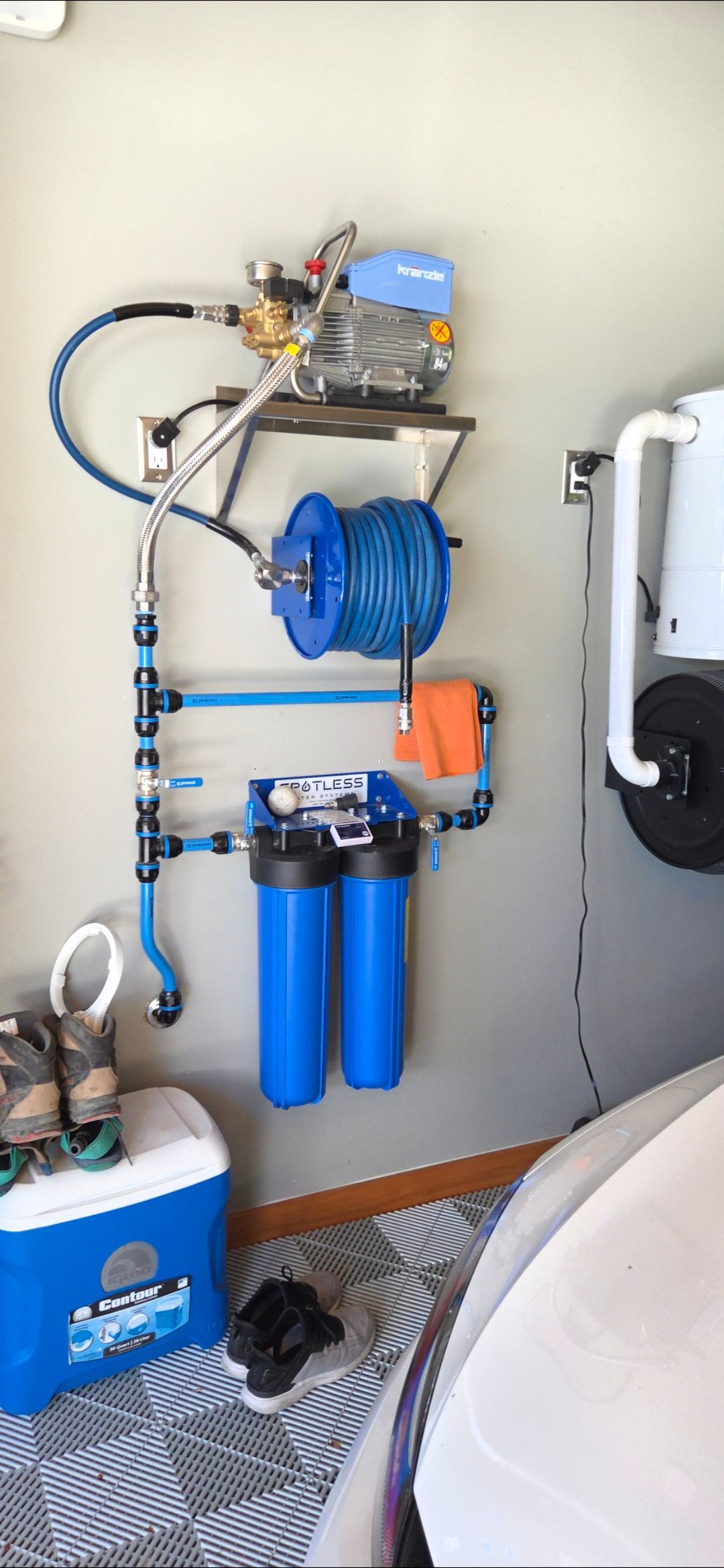 A wall-mounted pressure washer system with a hose reel, dual blue water filters, and plumbing, located in a garage.