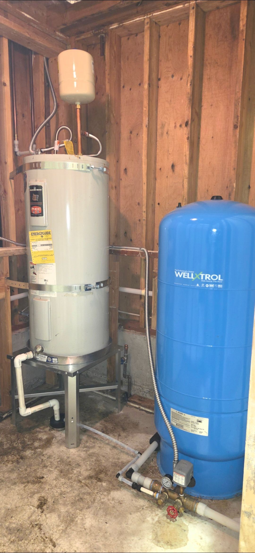 An electric water heater with an expansion tank above it sits next to a large blue well pressure tank in a utility room.