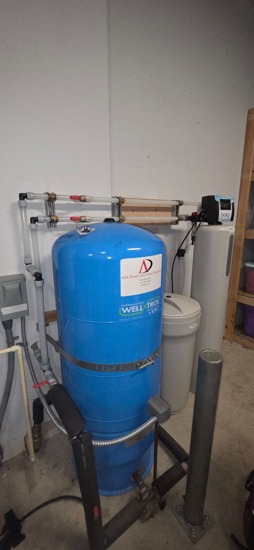 A water well pressure tank and filtration system with pipes and controllers installed against a white wall.