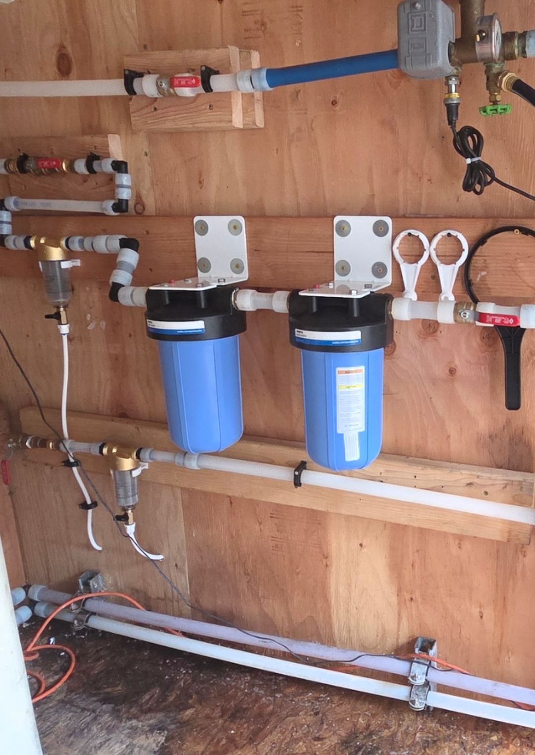 A dual-filter water filtration system mounted on a wooden wall, connected by white PEX tubing and plumbing fittings.