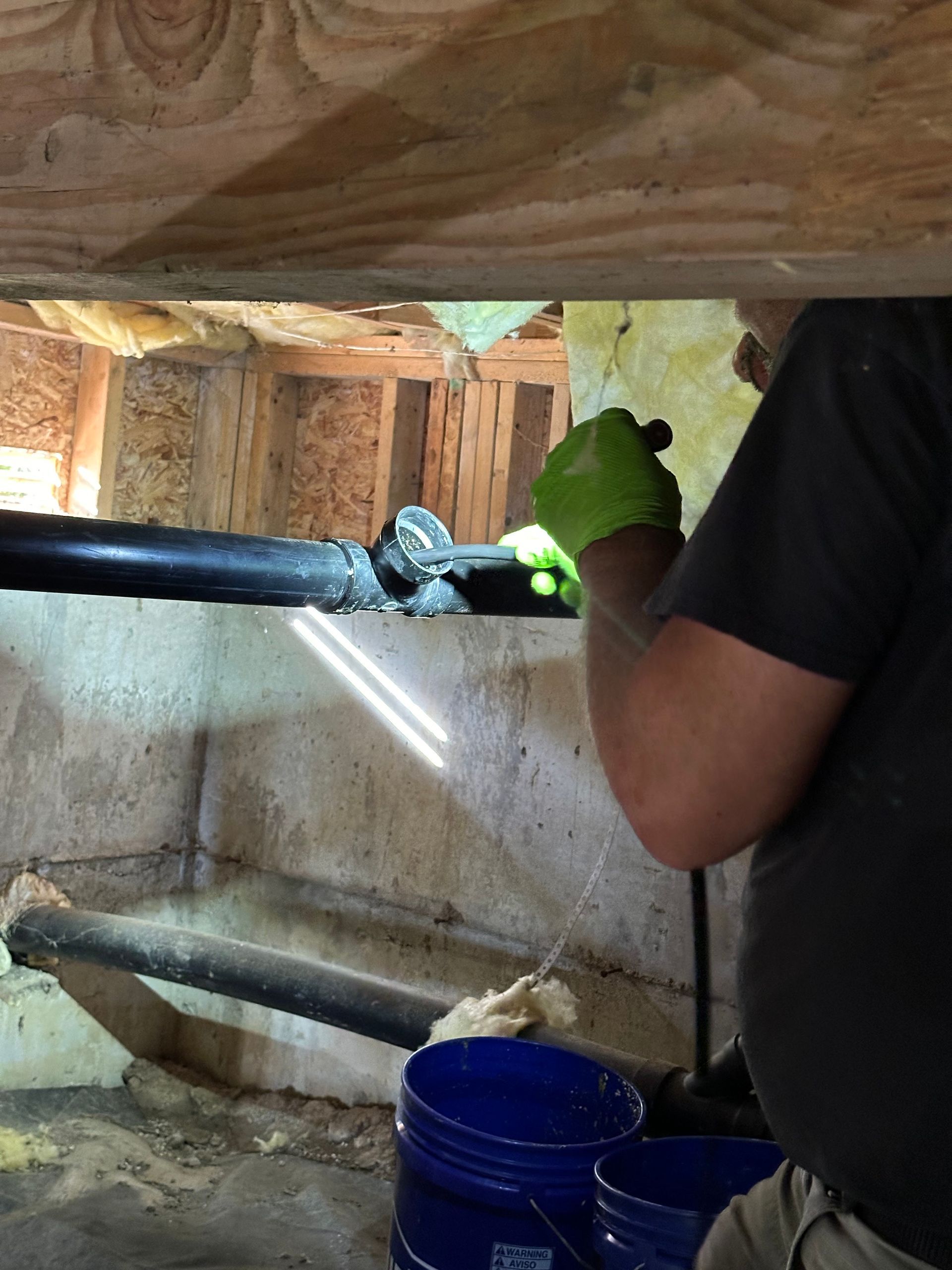 A person wearing green gloves uses a tool to work on a black drainage pipe in a crawlspace with blue buckets below.