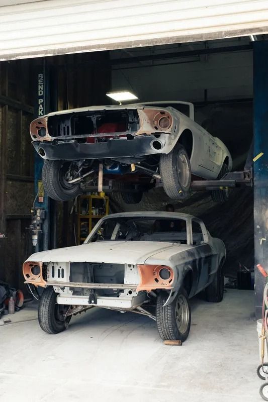 Two unfinished vintage cars stacked in a dim garage, one on a lift above the other. | Bob's Auto Repair