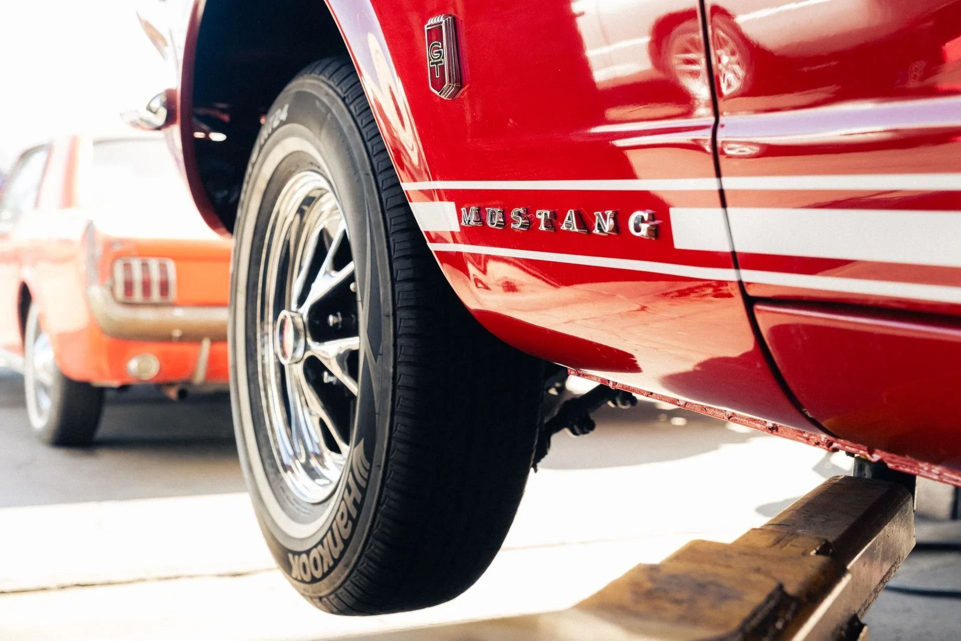 Red sports car on a lift, close-up of rear wheel and Mustang logo, orange car in background | Bob's Auto Repair
