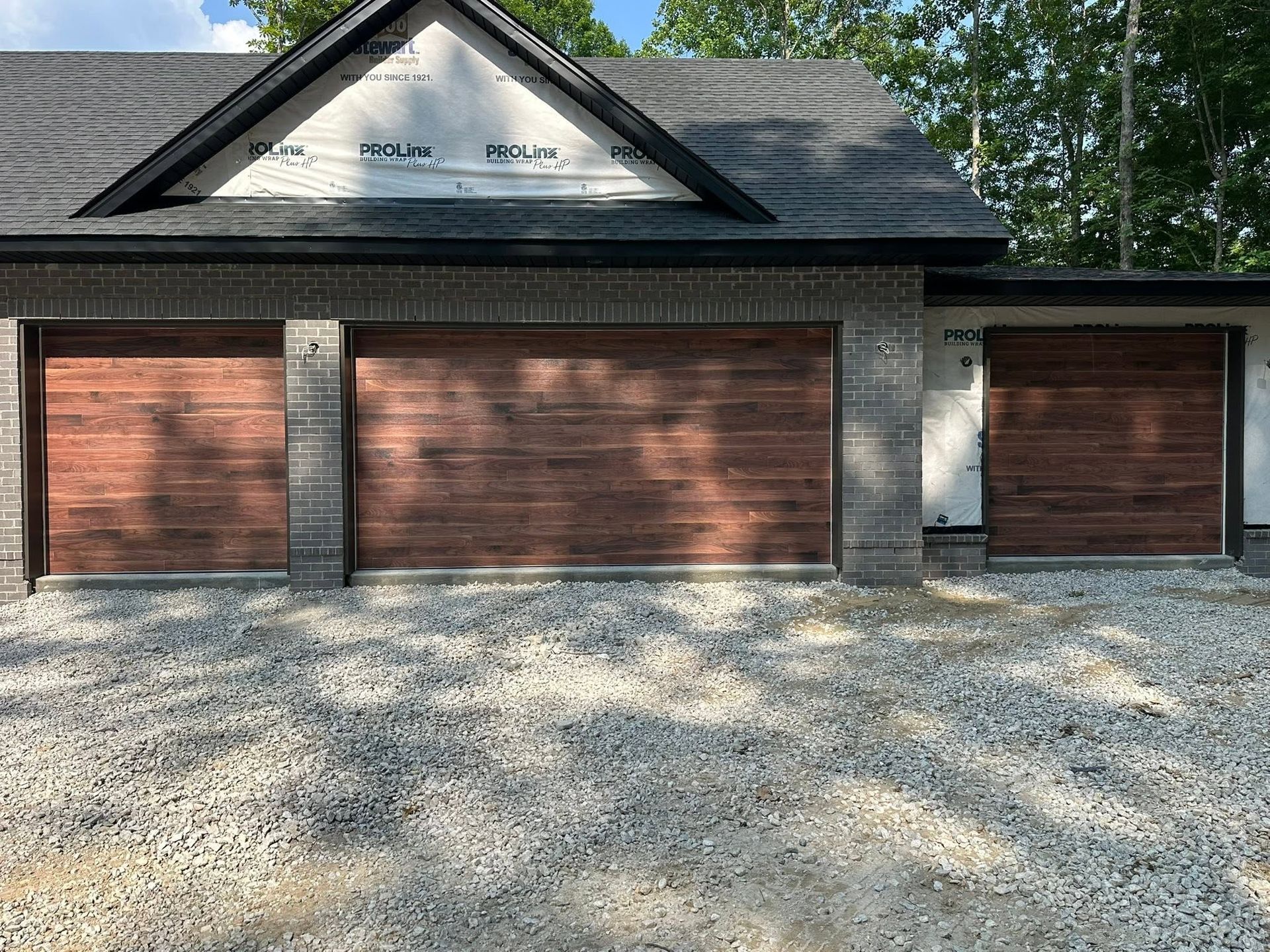 Three brown garage doors on a gray gravel driveway beneath a dark shingled roof. Three brown garage doors on a gray gravel driveway beneath a dark shingled roof.