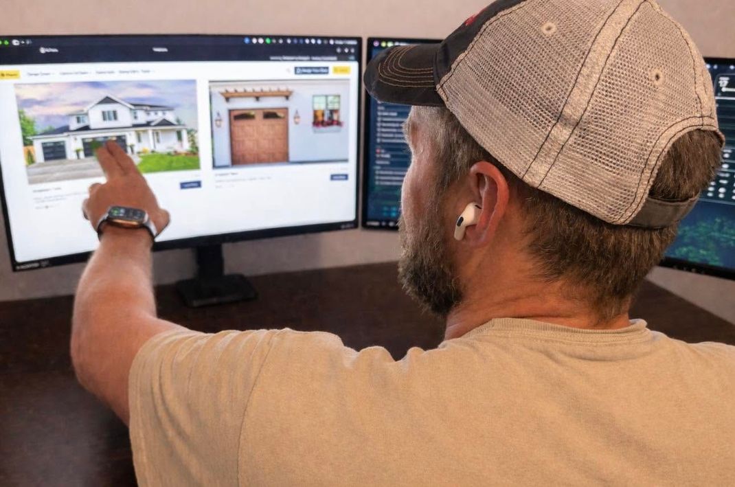 Person in a cap points at home listings on dual computer monitors in an office setting
