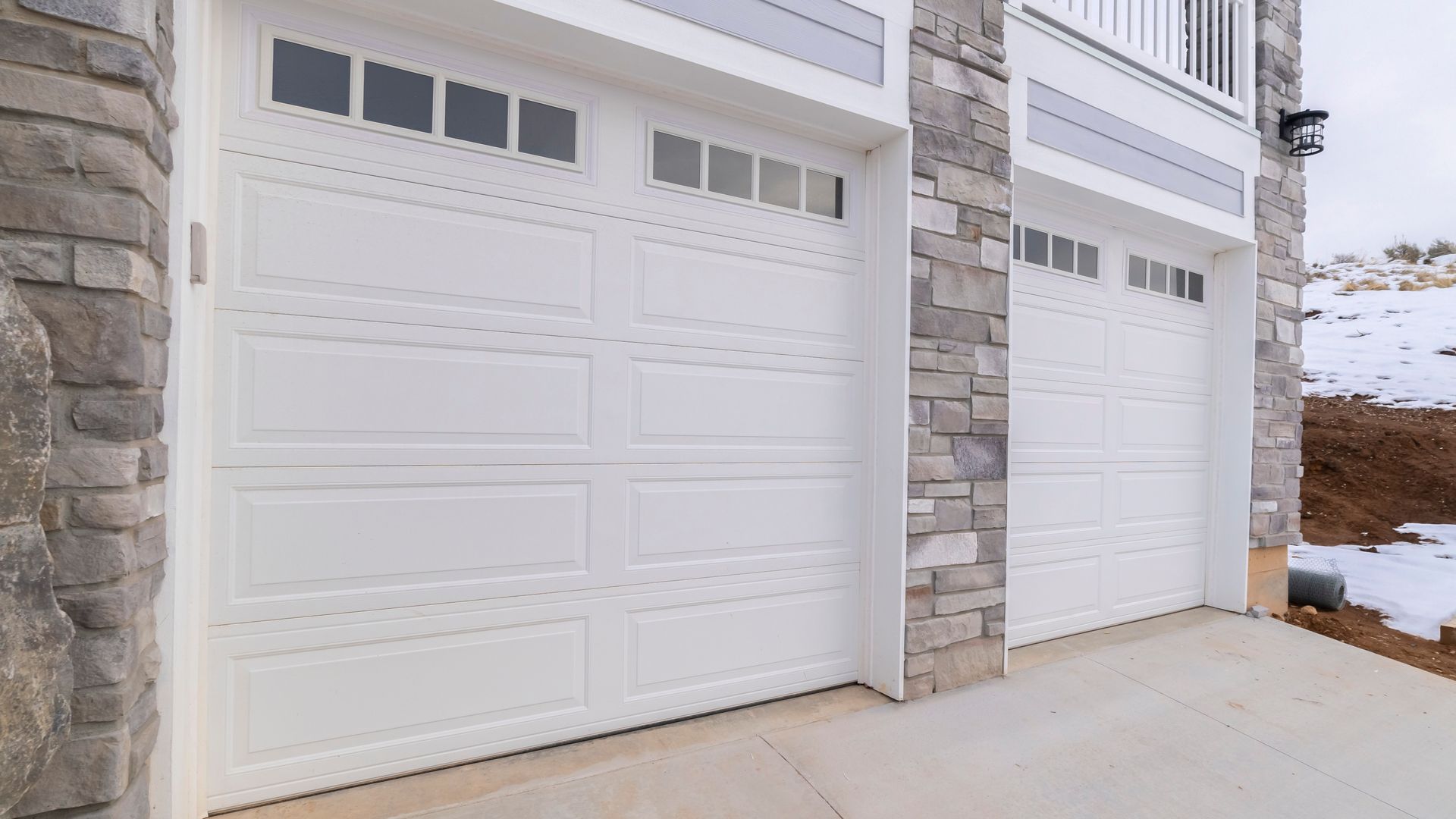 Two white garage doors on a stone-faced house beside a snowy driveway