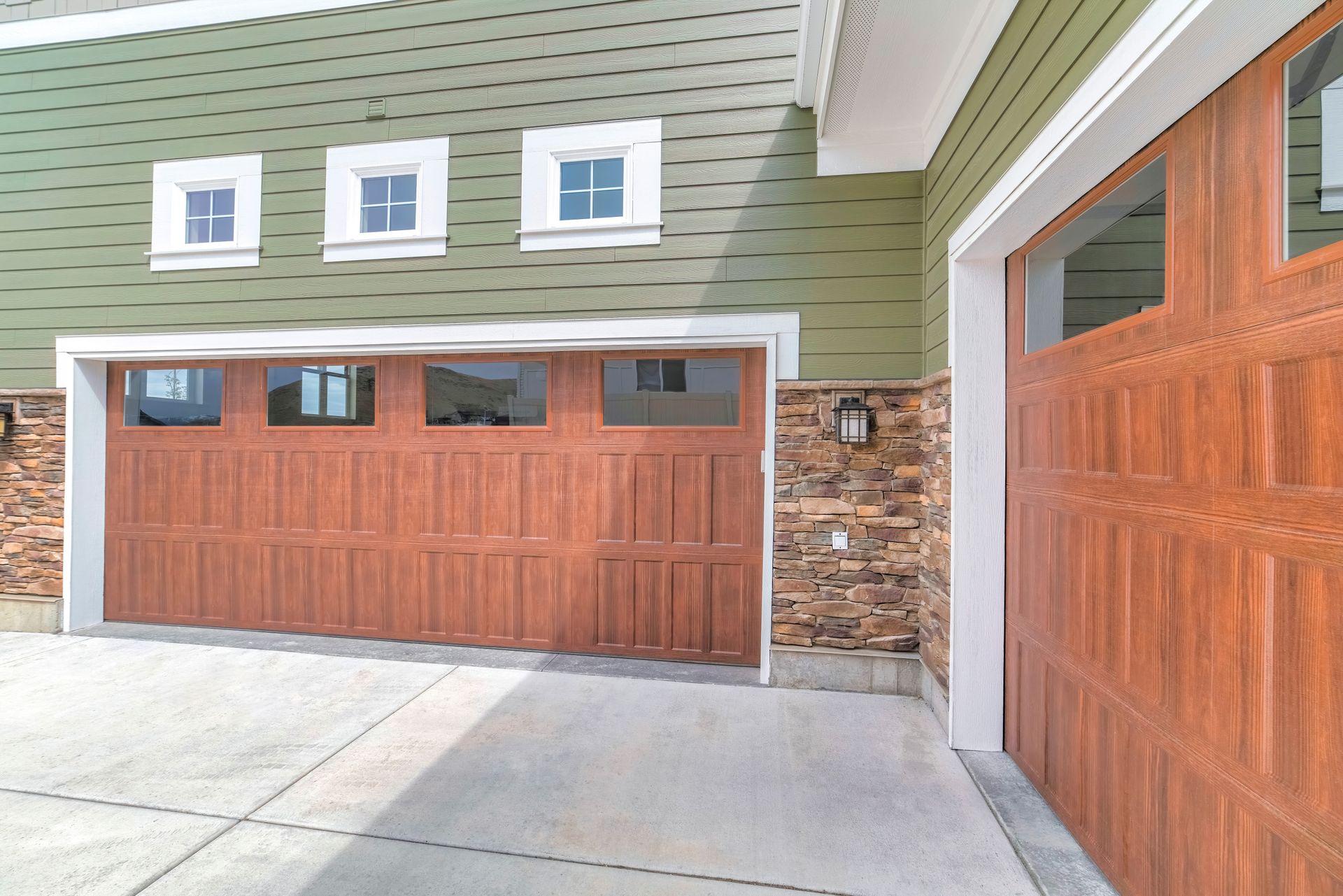Wooden two-car garage door beneath a teal house exterior, with gravel driveway in front. Wooden two-car garage door beneath a teal house exterior, with gravel driveway in front.
