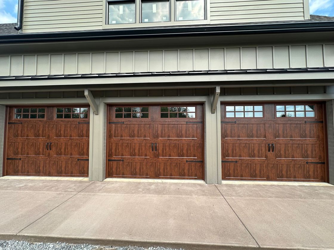 Two brown wooden garage doors in a brick building, with sunlight and shadows across the driveway.