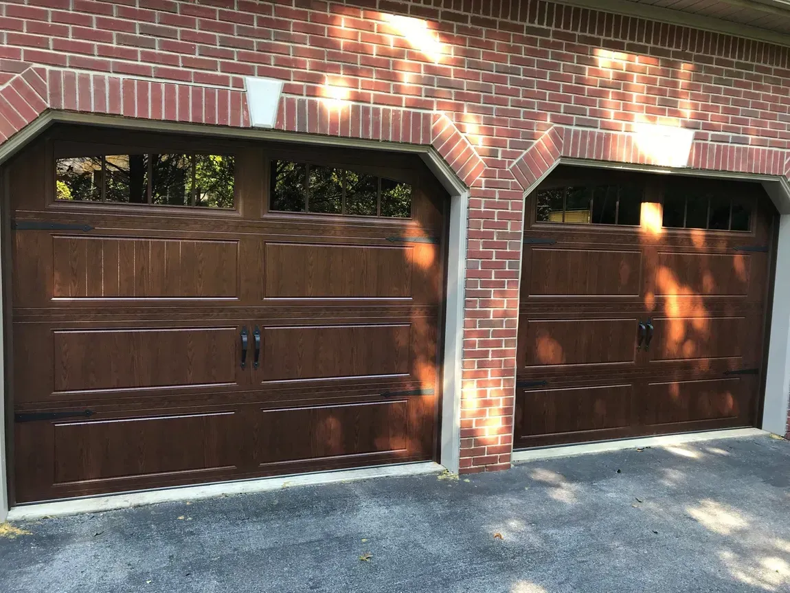 Two brown wooden garage doors in a brick building, with sunlight and shadows across the driveway.