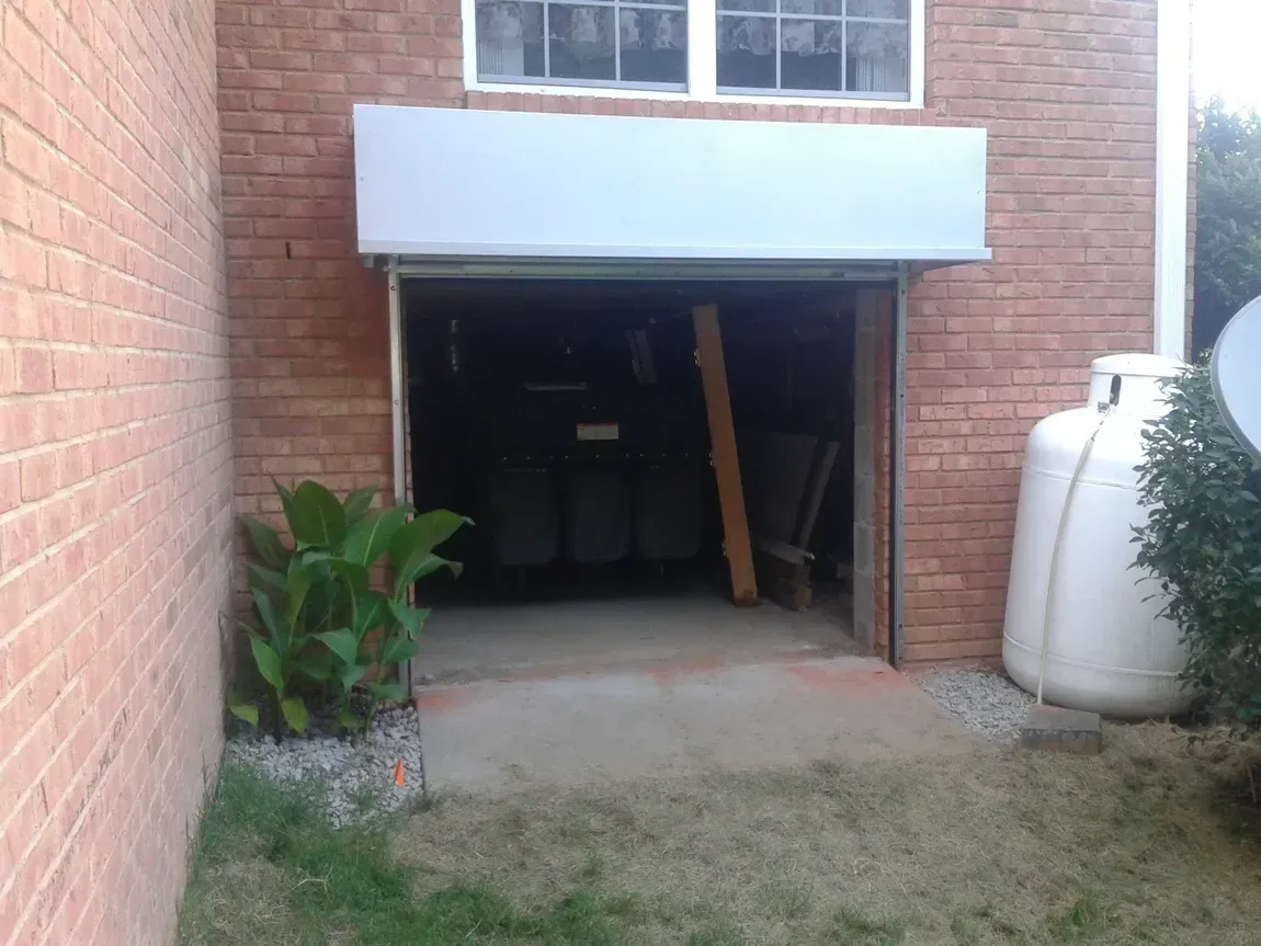 Brick house basement entrance with open garage-style door, lawn, potted plant, and large white tank beside it