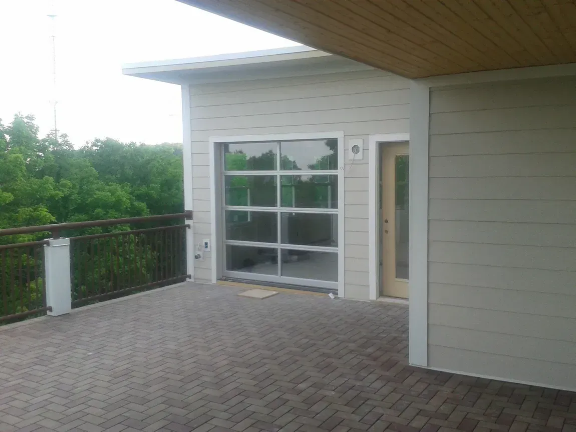 Outdoor patio with brick pavers, glass railing, and a modern house exterior under an overhang