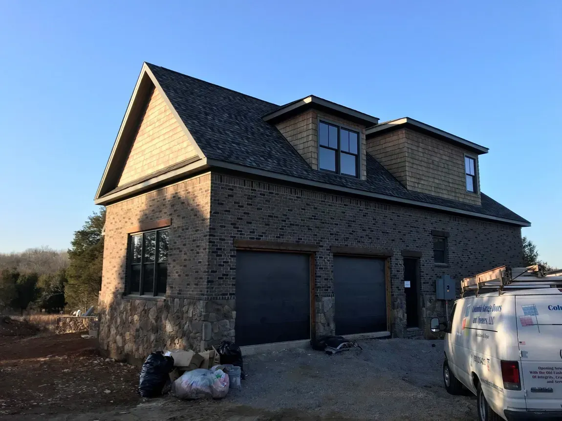 Stone house with dark roof, garage doors, and a white truck parked beside it under a clear blue sky