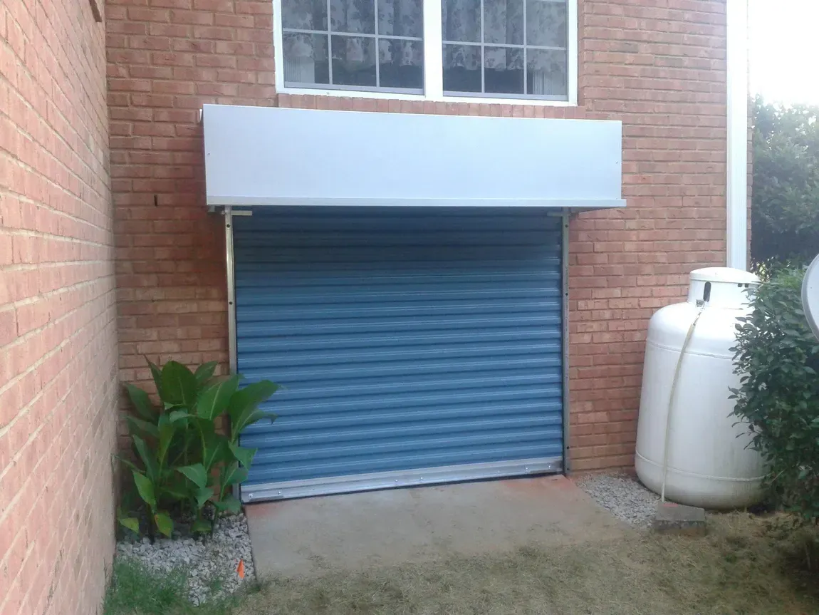 Blue roll-up garage door on a brick building, with a white tank and potted plants beside it.