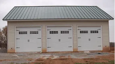 Three-car detached garage with white doors and a green metal roof on a bare lot