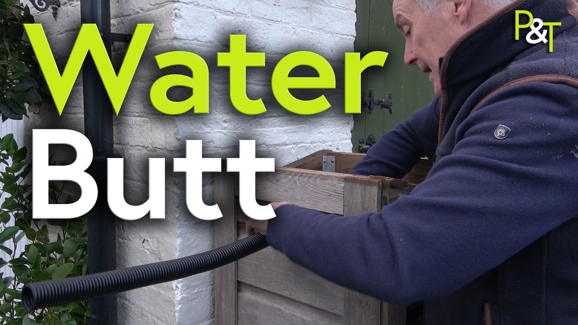 Martin Fish seen from behind, is left of frame. He is pushing a black pipe into the wooden cover of a water butt which is placed against a white painted brick wall. The text Water Butt in green and white over the top.
