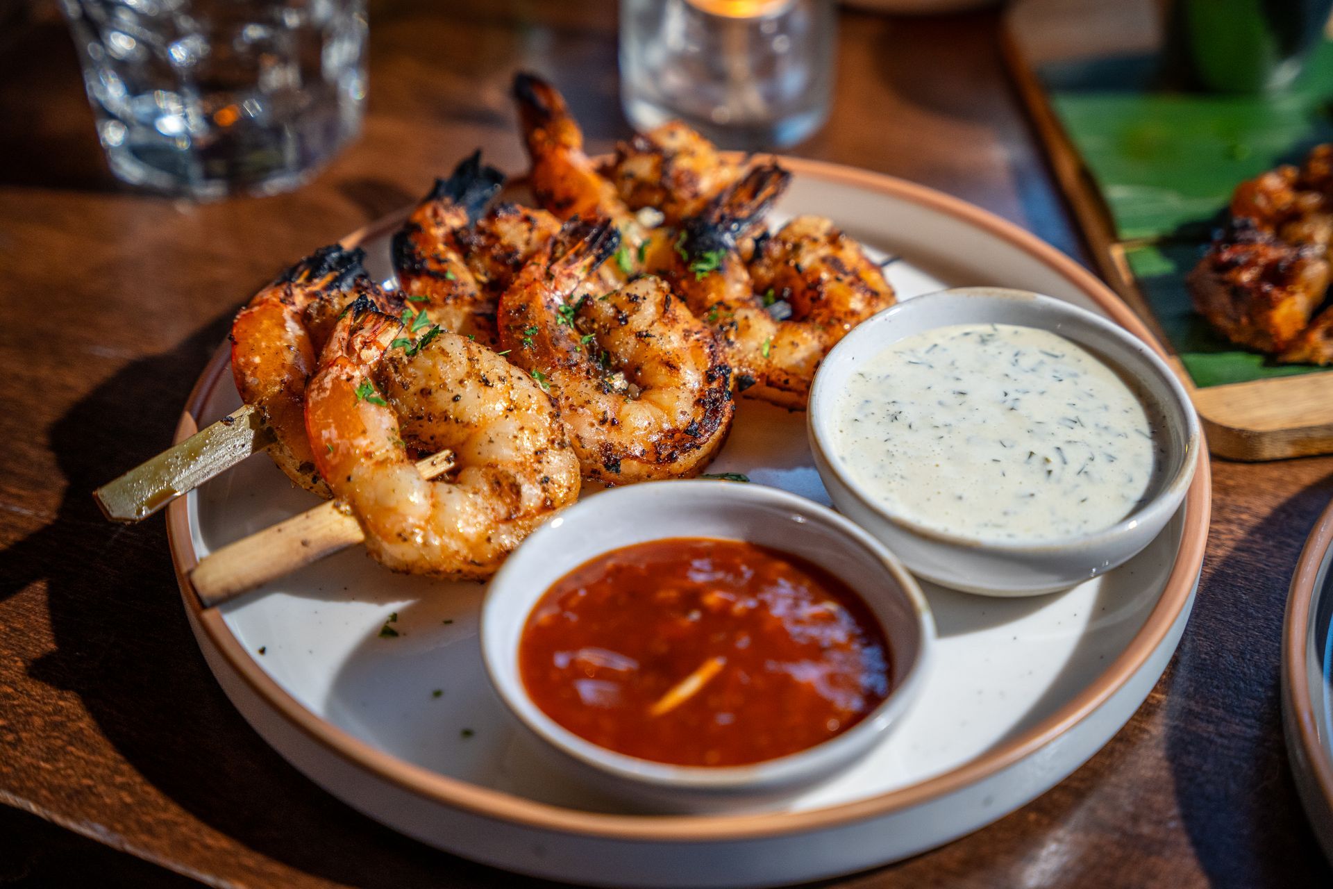 Grilled shrimp skewers with red and white dipping sauces on a plate in a restaurant setting