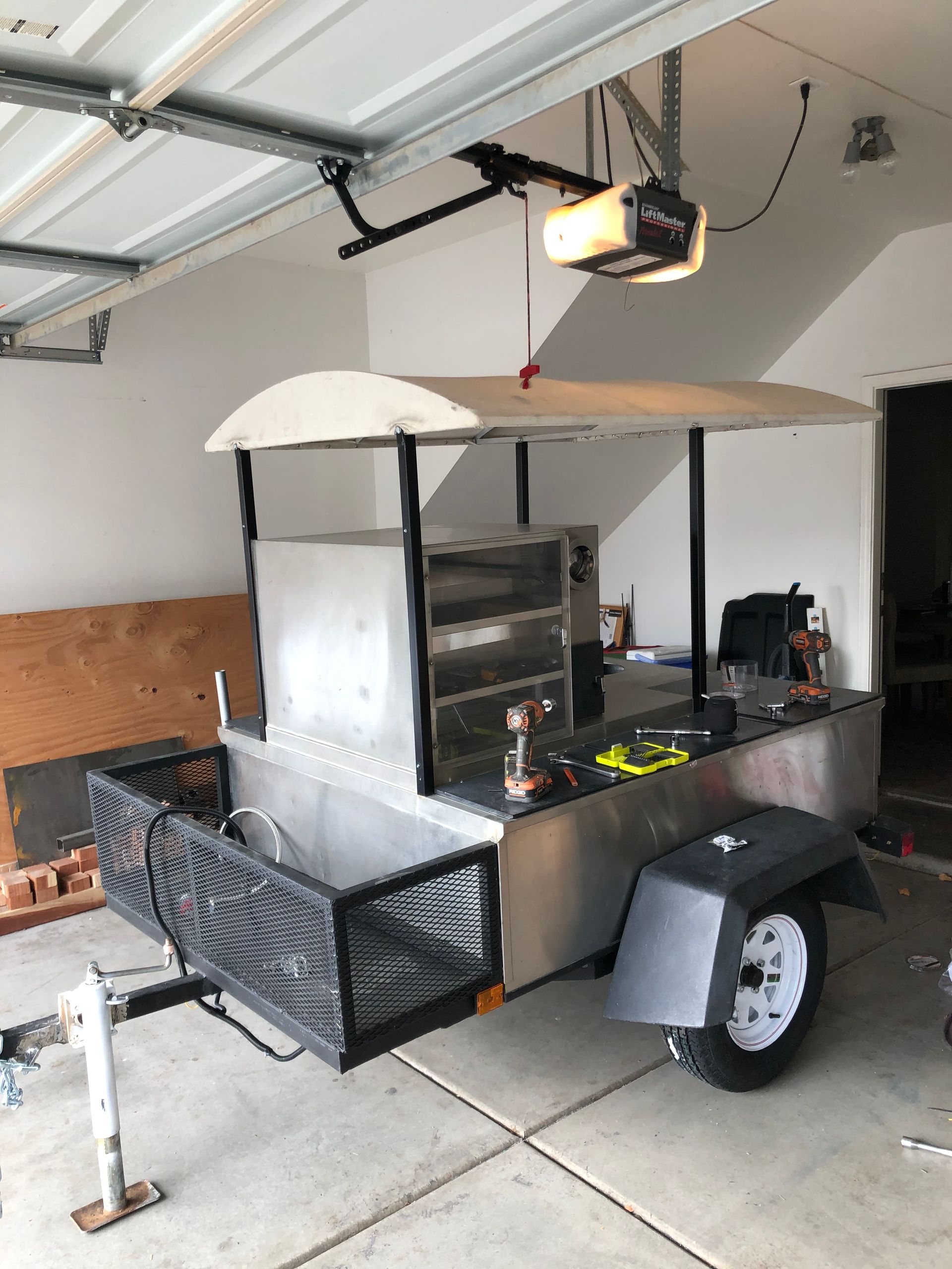 Black trailer-style mobile grill cart in a garage with a metal canopy and one wheel visible