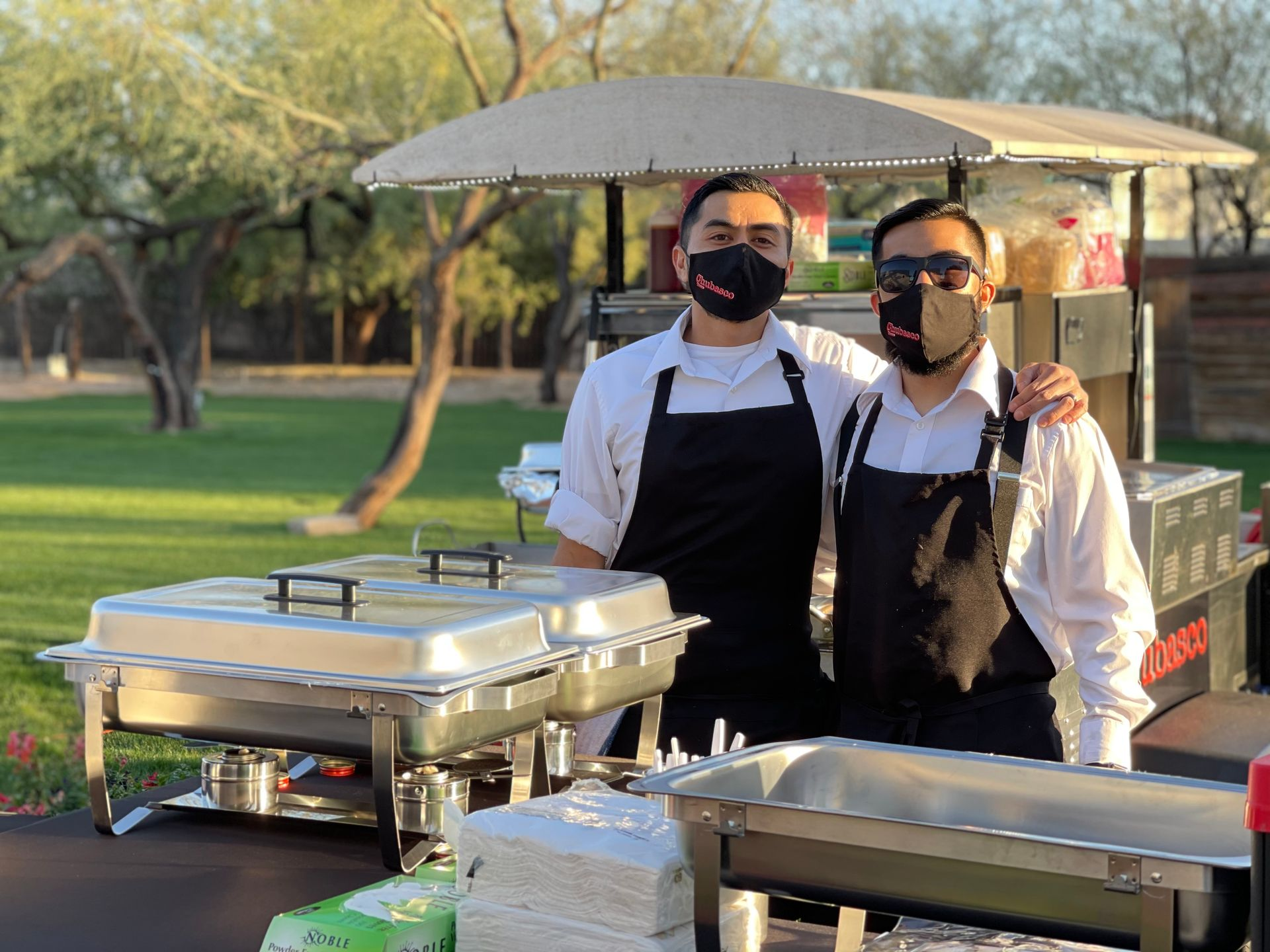 Two masked people in aprons standing behind buffet trays at an outdoor park event