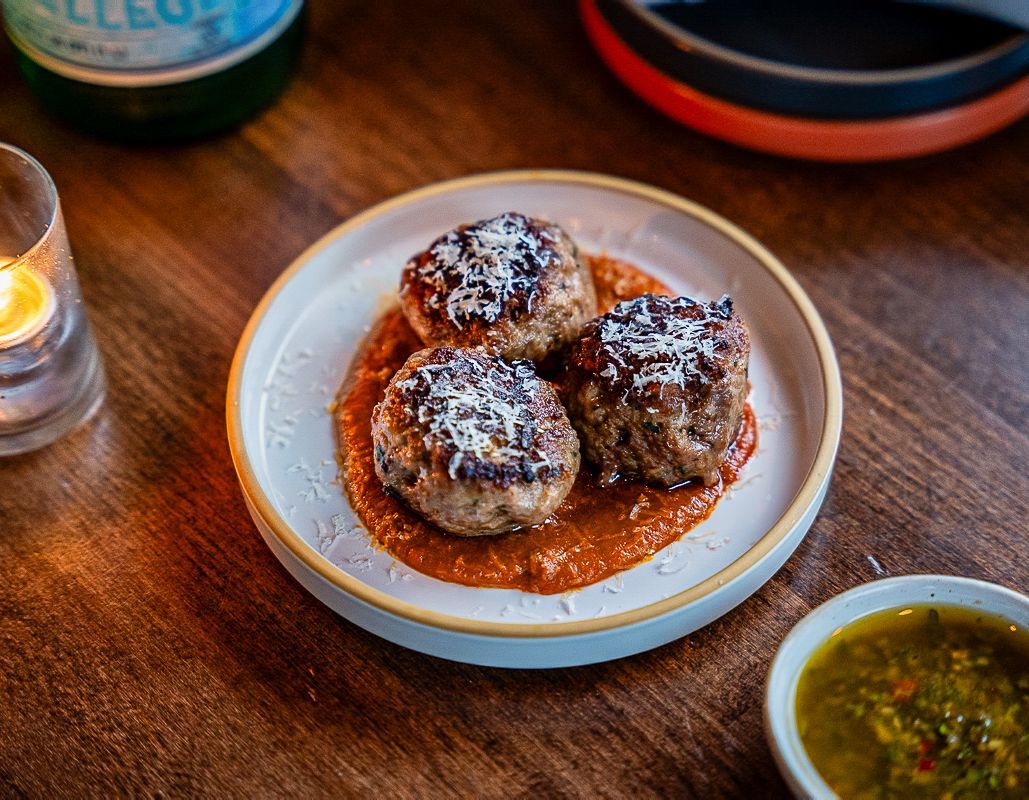 Three meatballs in red sauce on a white plate beside a small bowl of green sauce on a wooden table
