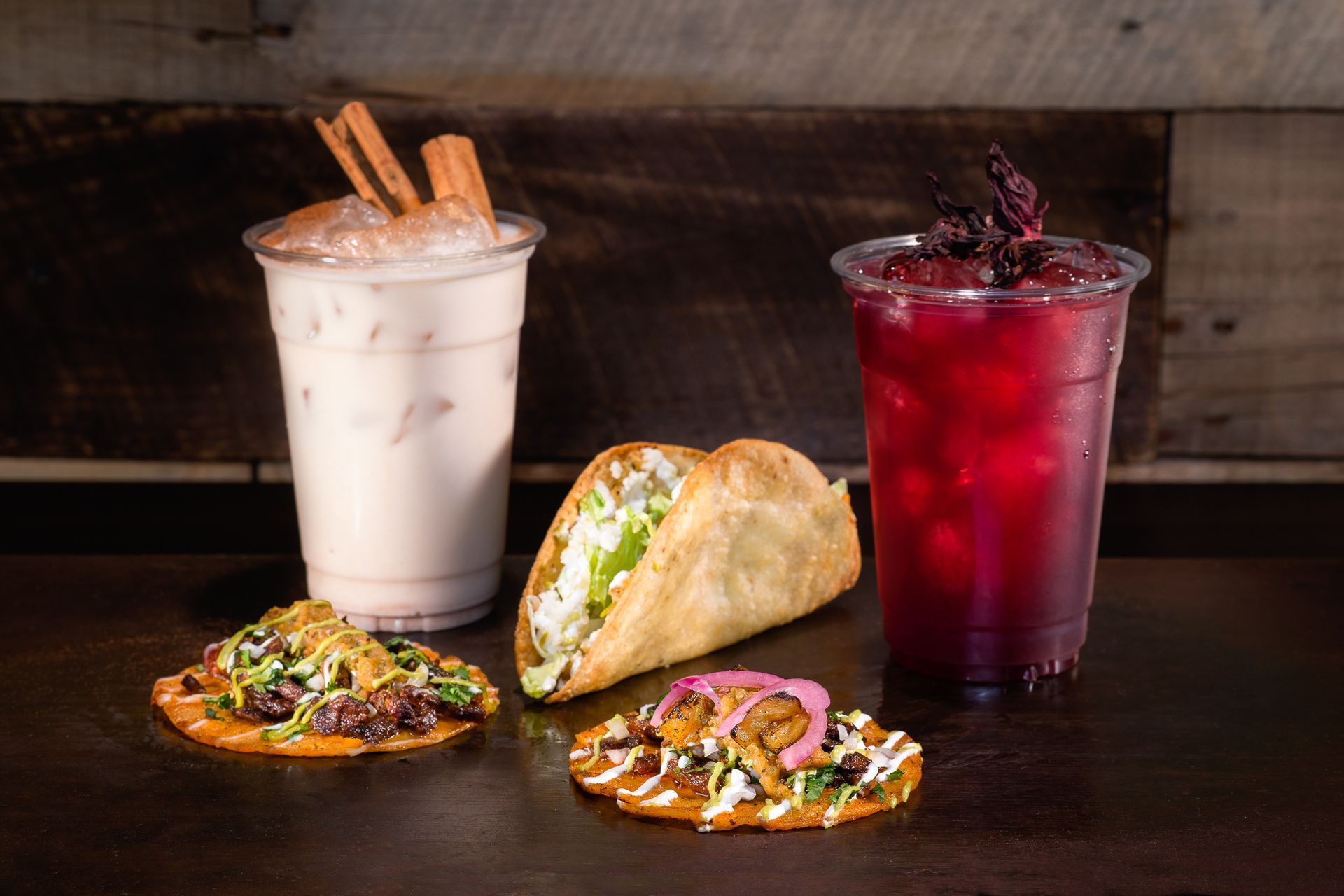Tacos and iced drinks on a dark table, with a white drink and a red drink in clear cups