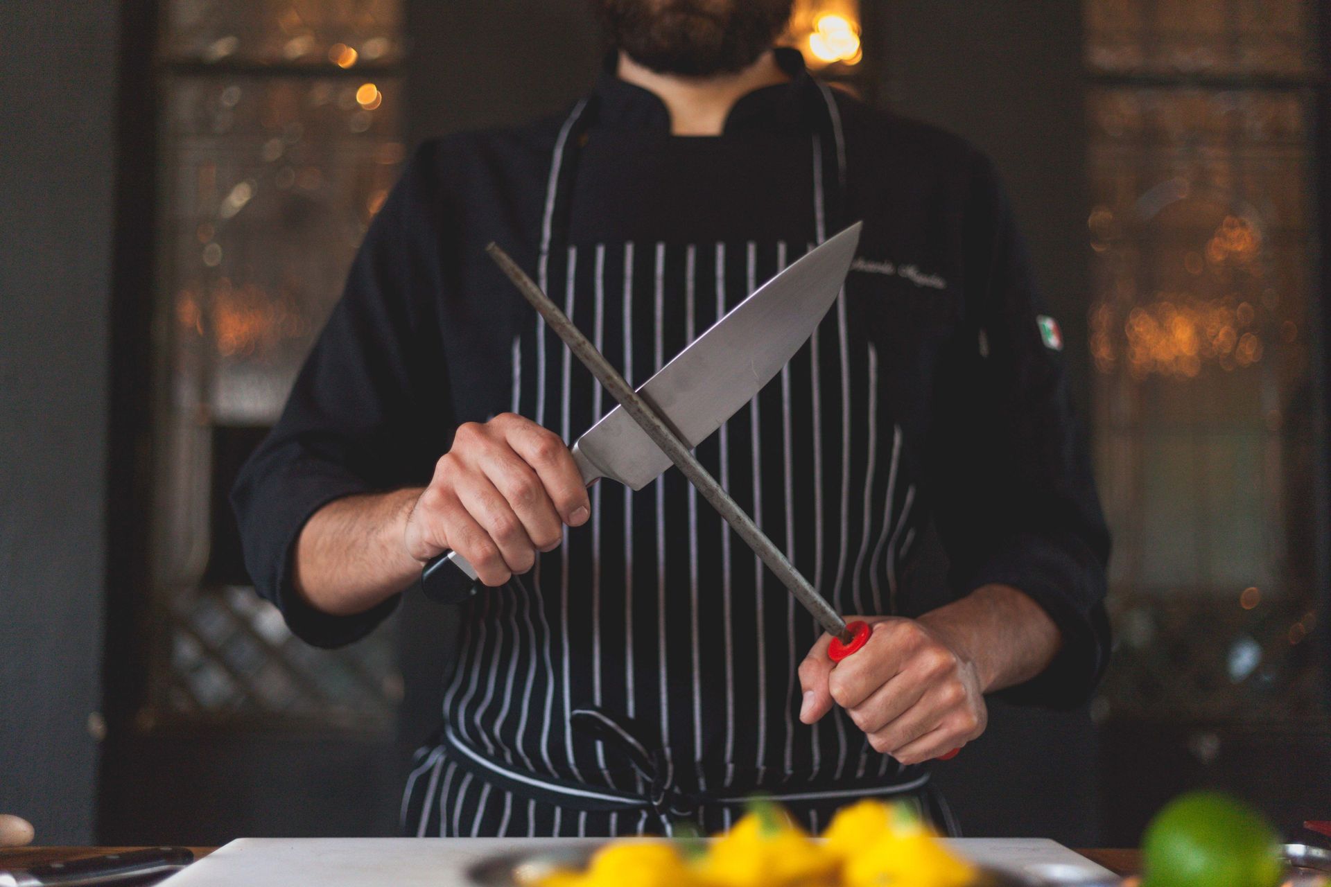 Chef sharpening a knife at a kitchen counter, wearing a black striped apron.
