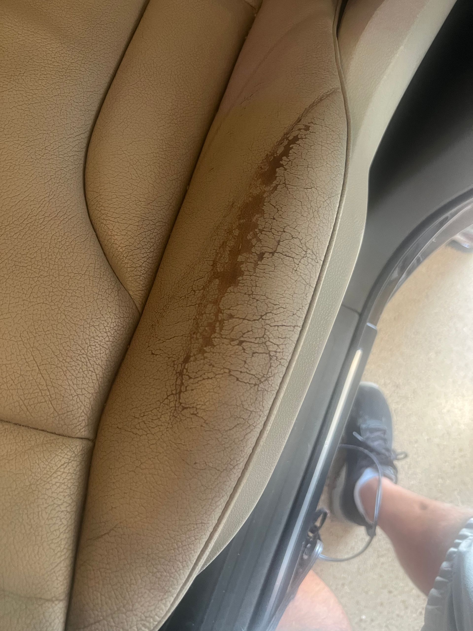 Close-up of a beige car seat with brown stains, next to a person's leg and shoe near the door.