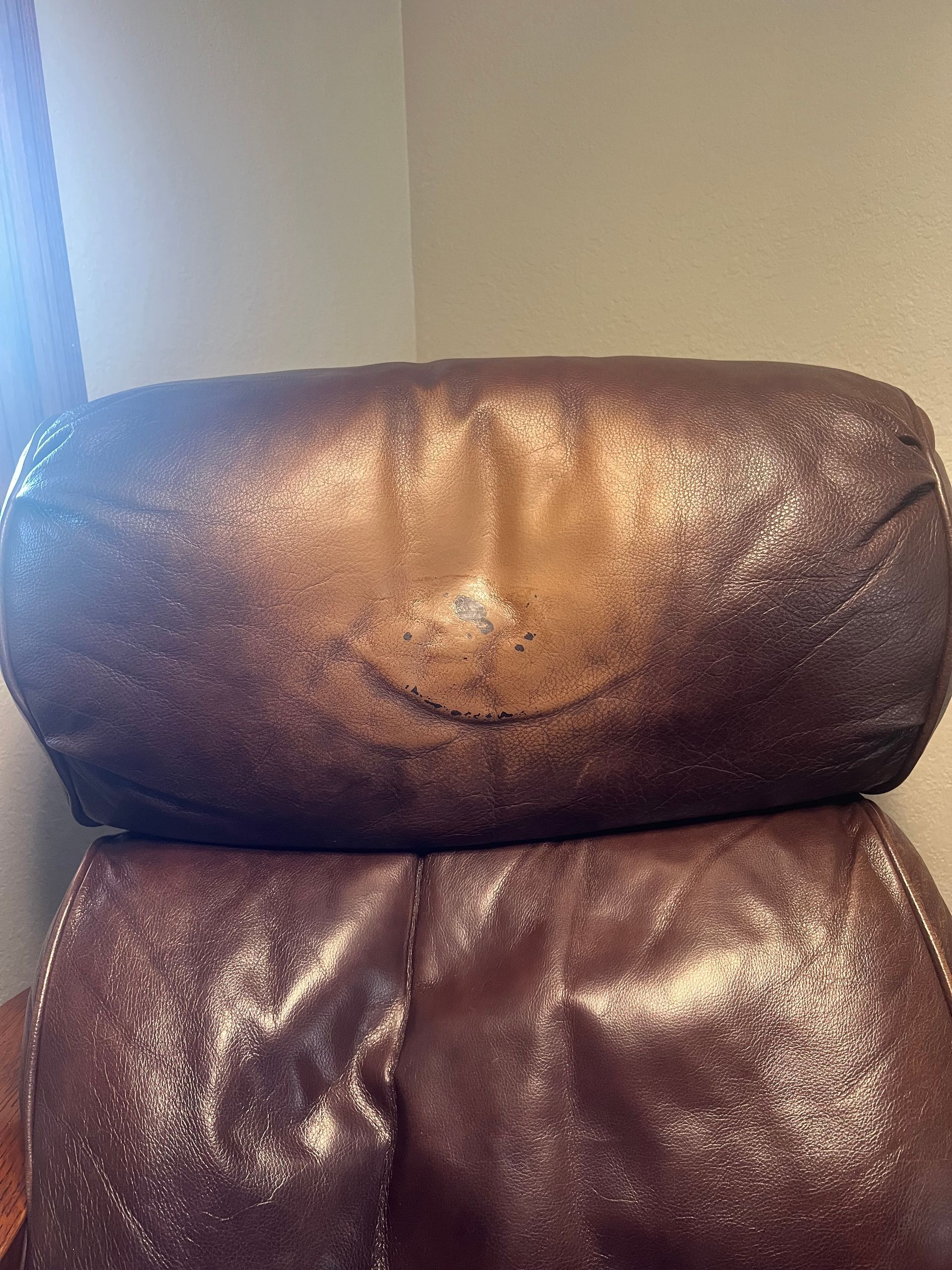 Brown leather chair back with a worn, cracked center seam against a light wall and window light.