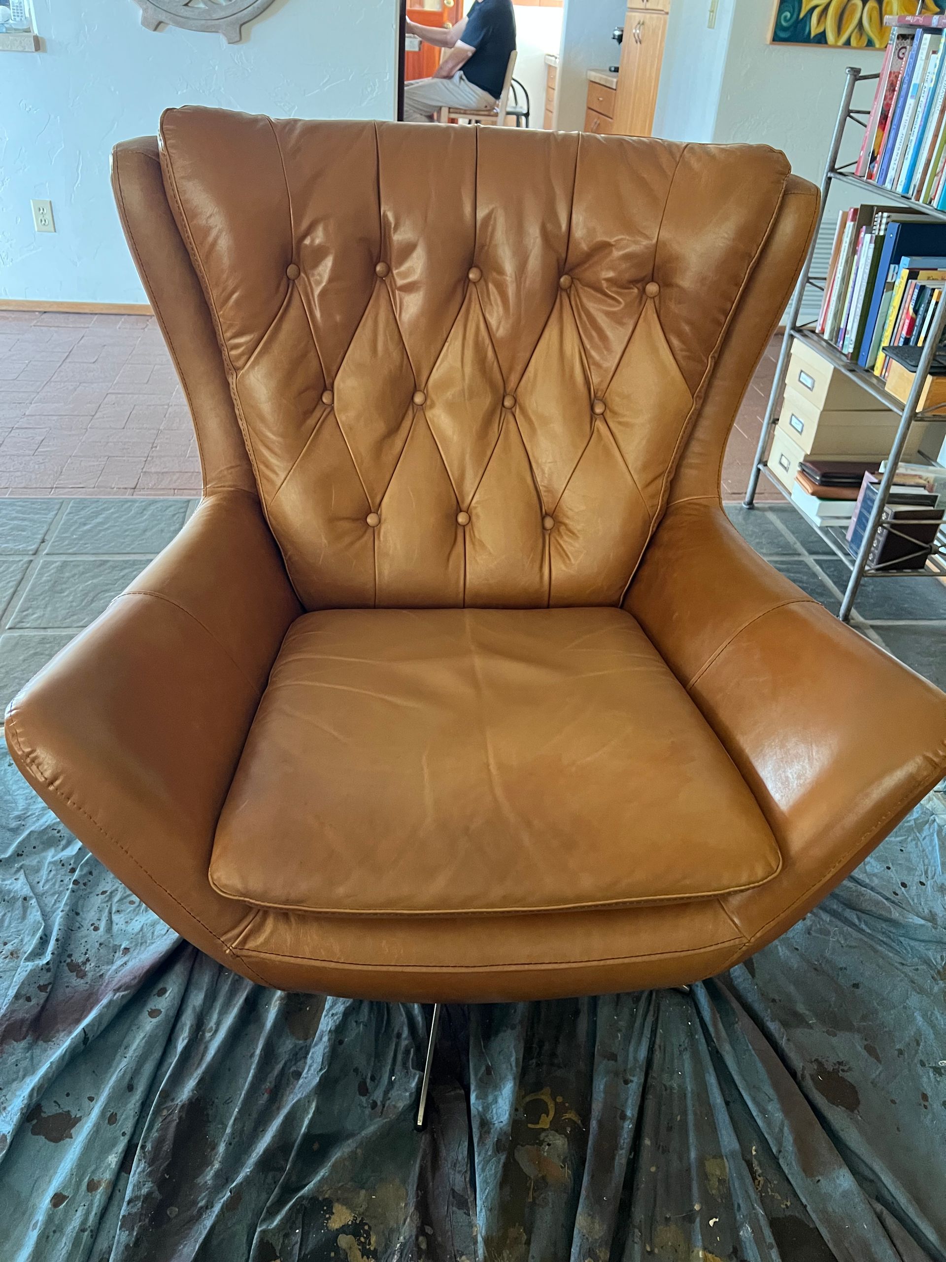 Brown tufted leather armchair with wide arms, sitting on a covered floor indoors.